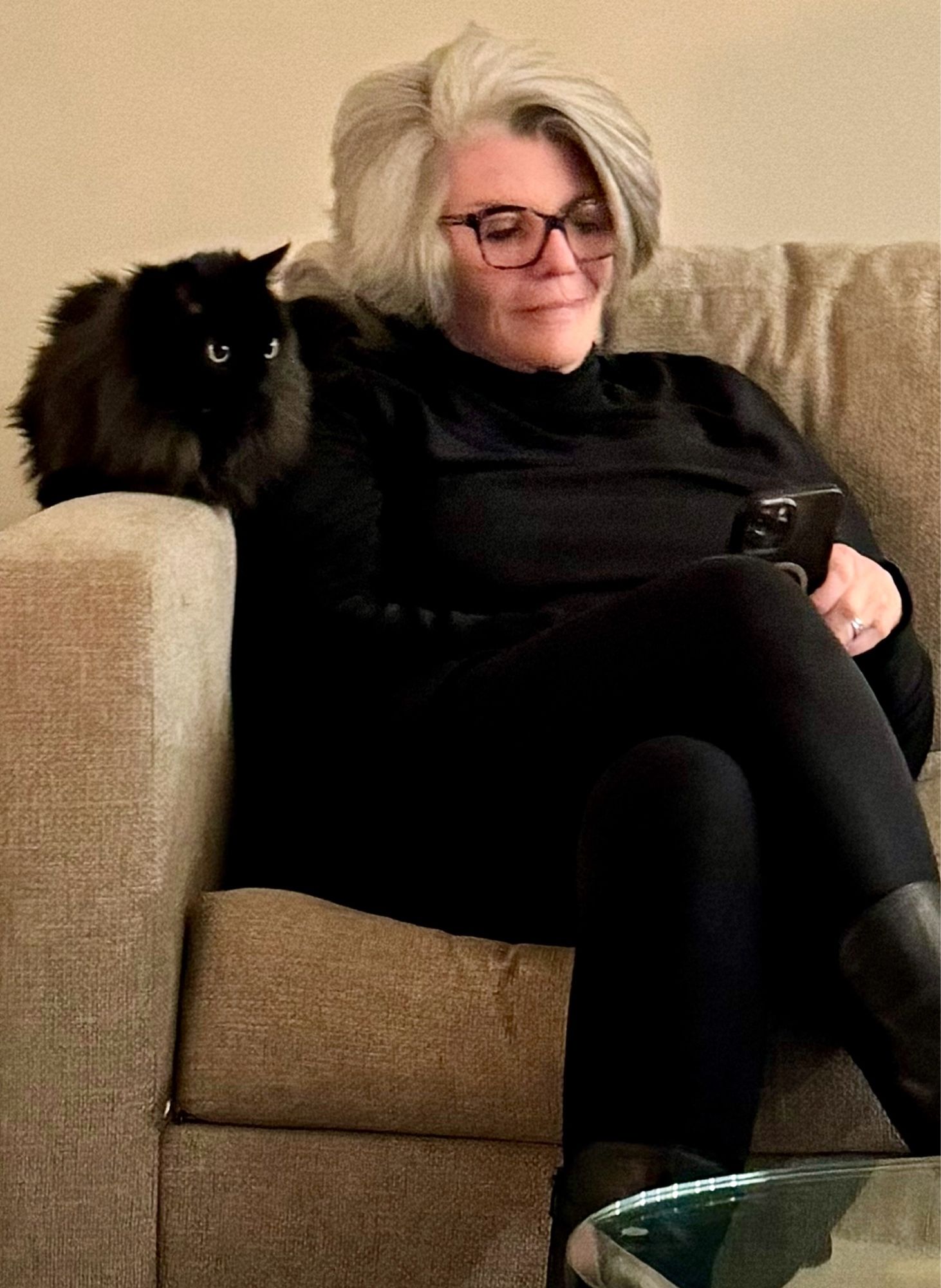 A photo of a long haired black cat with yellow eyes perched on the arm of a white sofa, sitting next to me.  I have silver hair, am wearing black clothes and have a black iPhone in my hand.