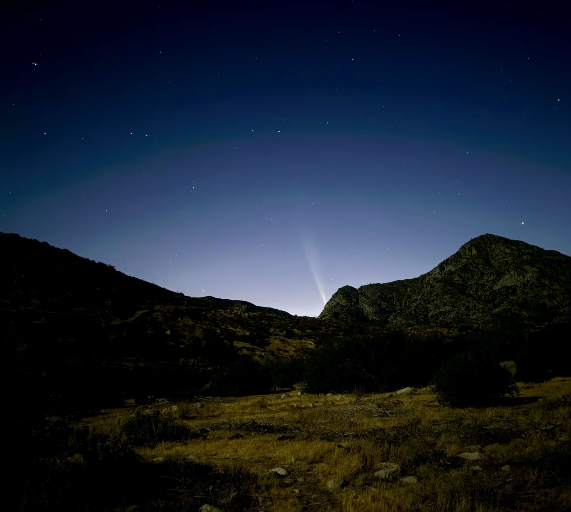 The faint tail of a comet is visible in the evening sky, along with a few stars.  In the foreground are mountains and a trail 