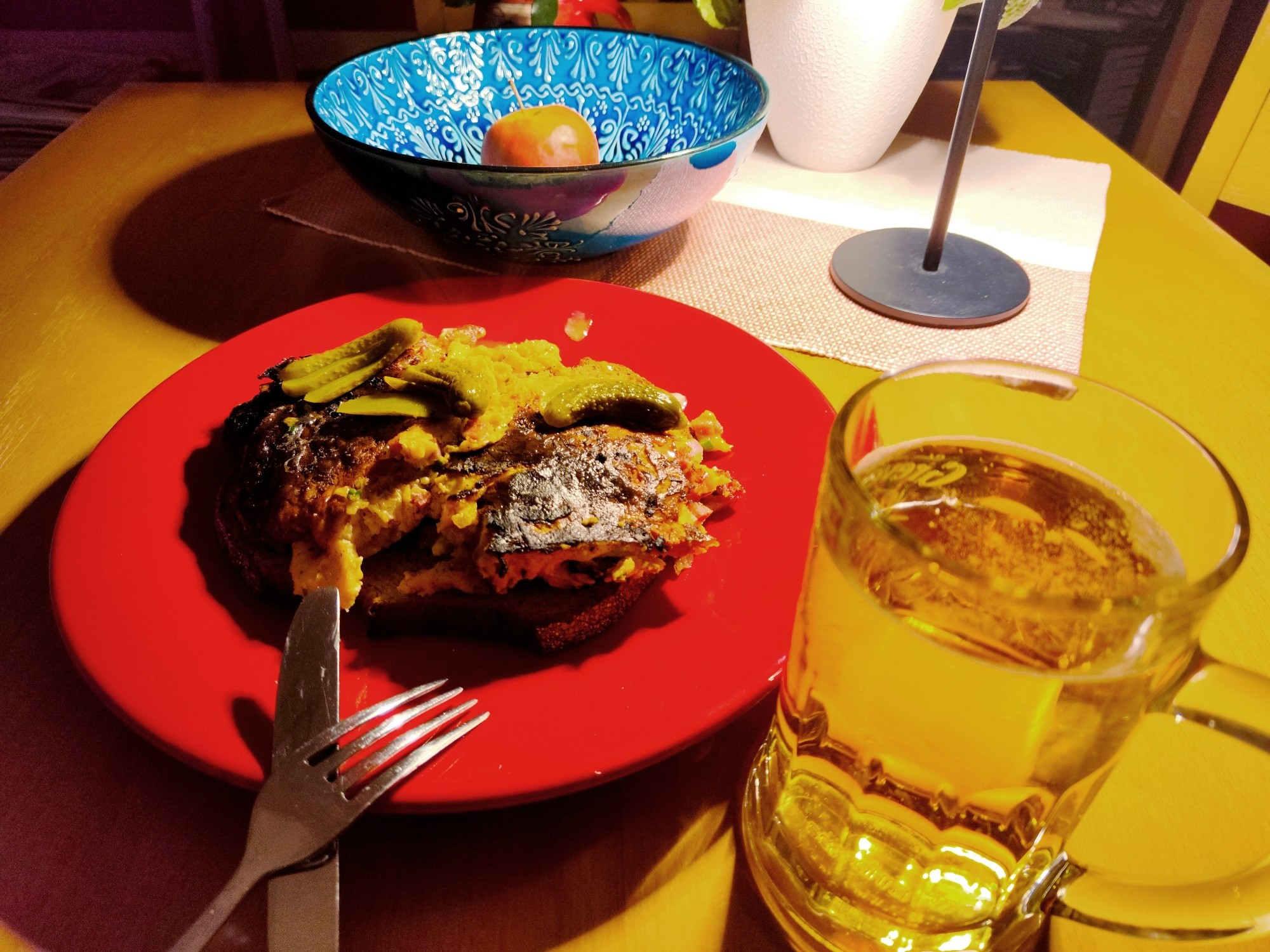 Omelette auf Brotscheiben auf einem roten Teller auf einem Holztisch, davor ein Glas Bier und Messer und Gabel, dahinter eine blaue Obstschale mit einem Apfel auf einem Tischdeckchen