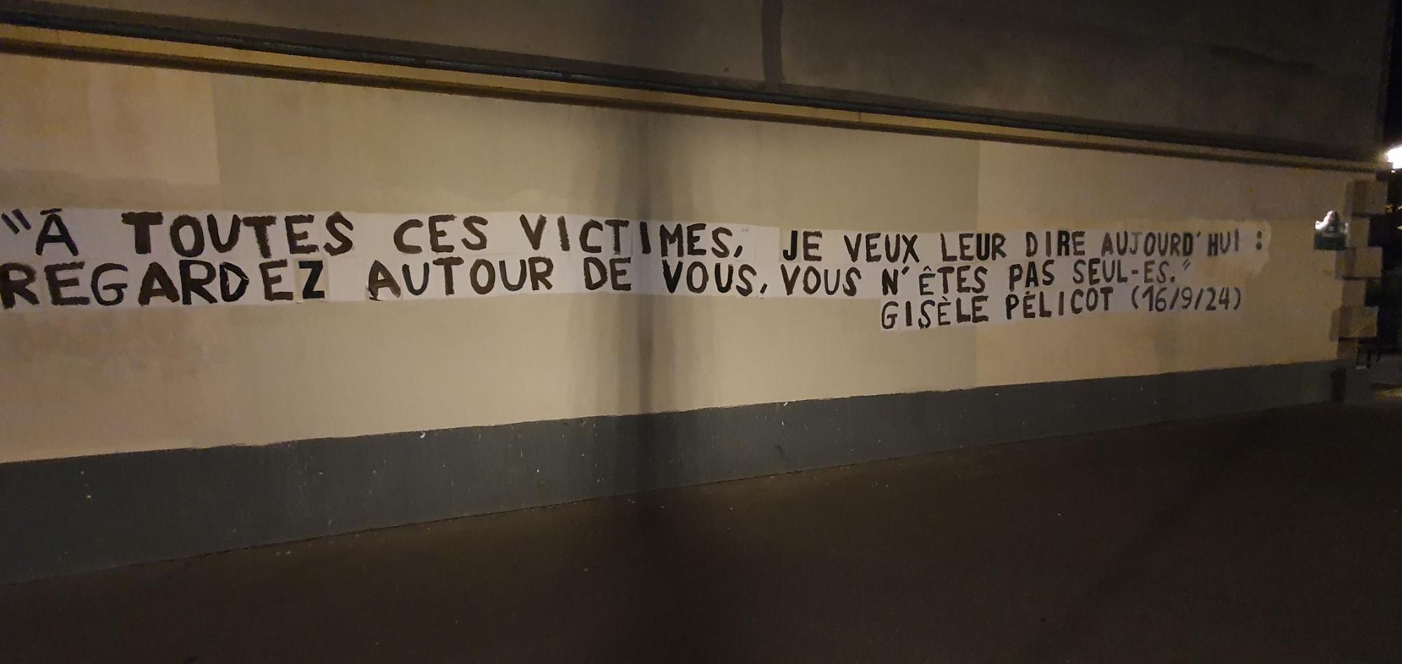 Foto einer weißen Wand mit schwarzer Aufschrift:

“À toutes ces victimes, je veux leur dire aujourd’hui : regardez autour de vous, vous n’êtes pas seuls” / 

Maschinell ins Deutsche übersetzt: „All diesen Opfern möchte ich heute sagen: Schaut euch um, ihr seid nicht allein“