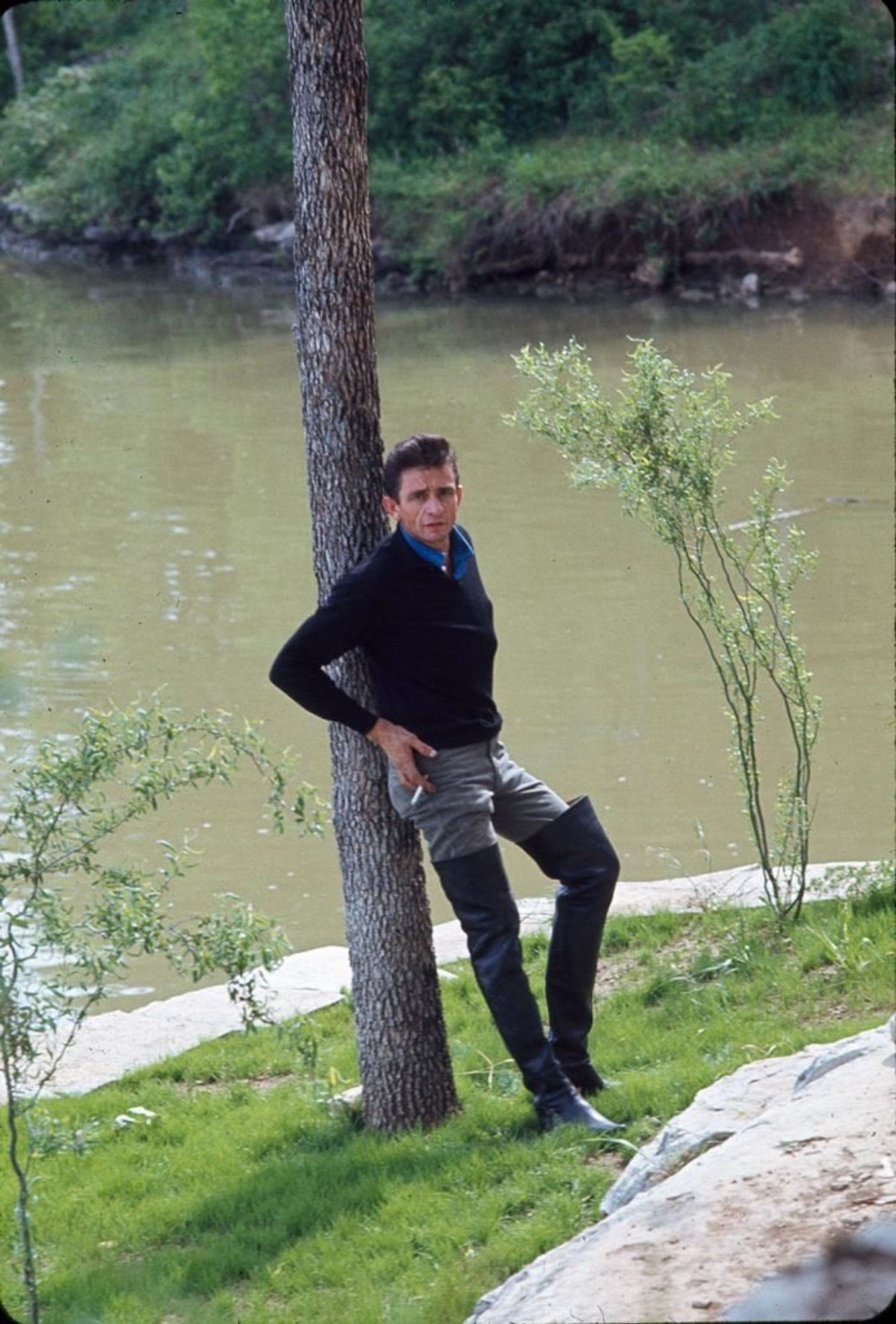 Singer Johnny Cash leaning nonchalantly on a tree next to a river, wearing a black sweater, tan pants, and black leather thigh-high boots.