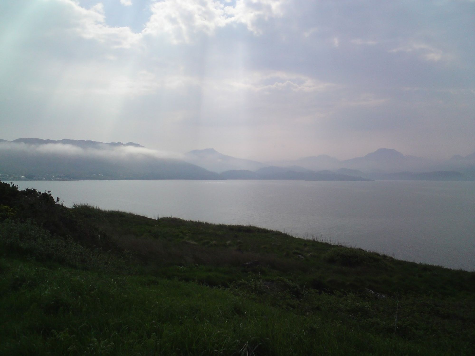 View of the sea and Torridon hills behind. Some thin low clouds/mist over the hills, some tops emerging. Cloud cover higher in the sky is pierced by some bright sunshine rays, with a little bit of blue sky visible to the top.