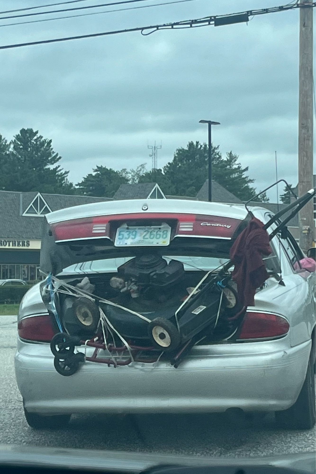 Car driving with trunk open and a lawn mower and bunch of other crap hanging out but not tied down.