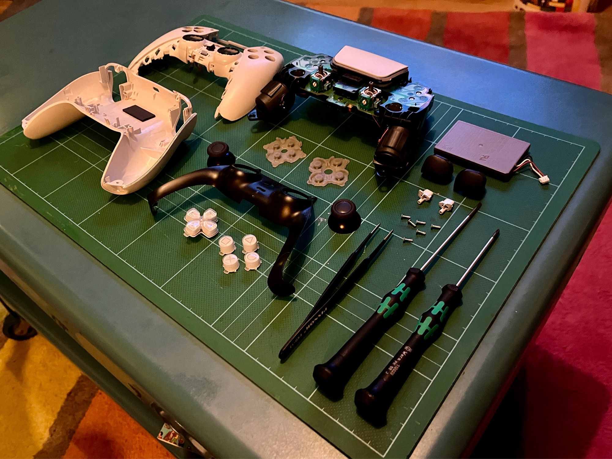 A complete PlayStation 5 DualSense controller’s parts, separated and neatly spread out on a deep green cutting mat with some screwdrivers and tweezers.