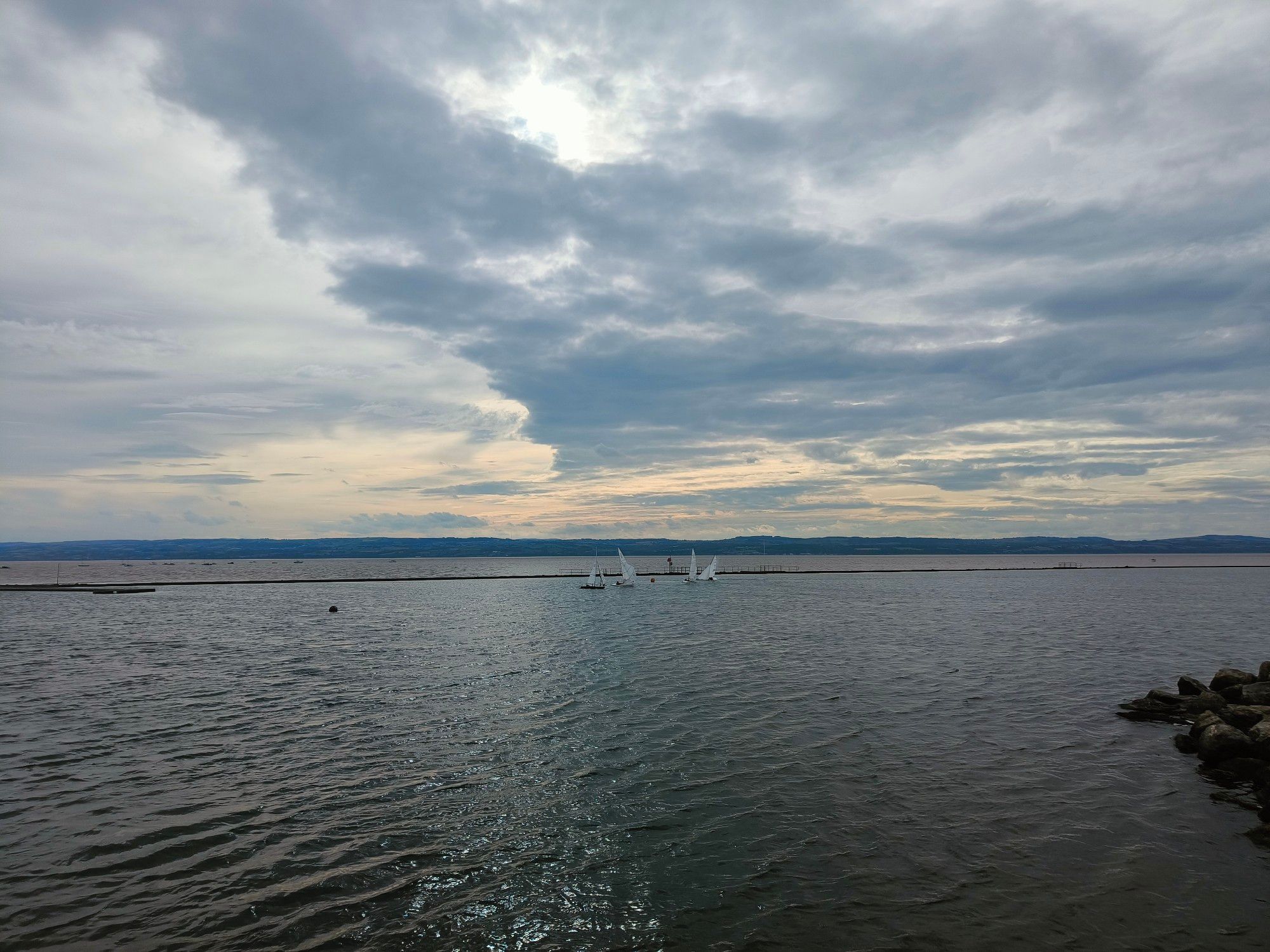 A cloudy sky with lowering sun light. In the background the low hills. In the foreground a marine lake with small sailing dinghies in view