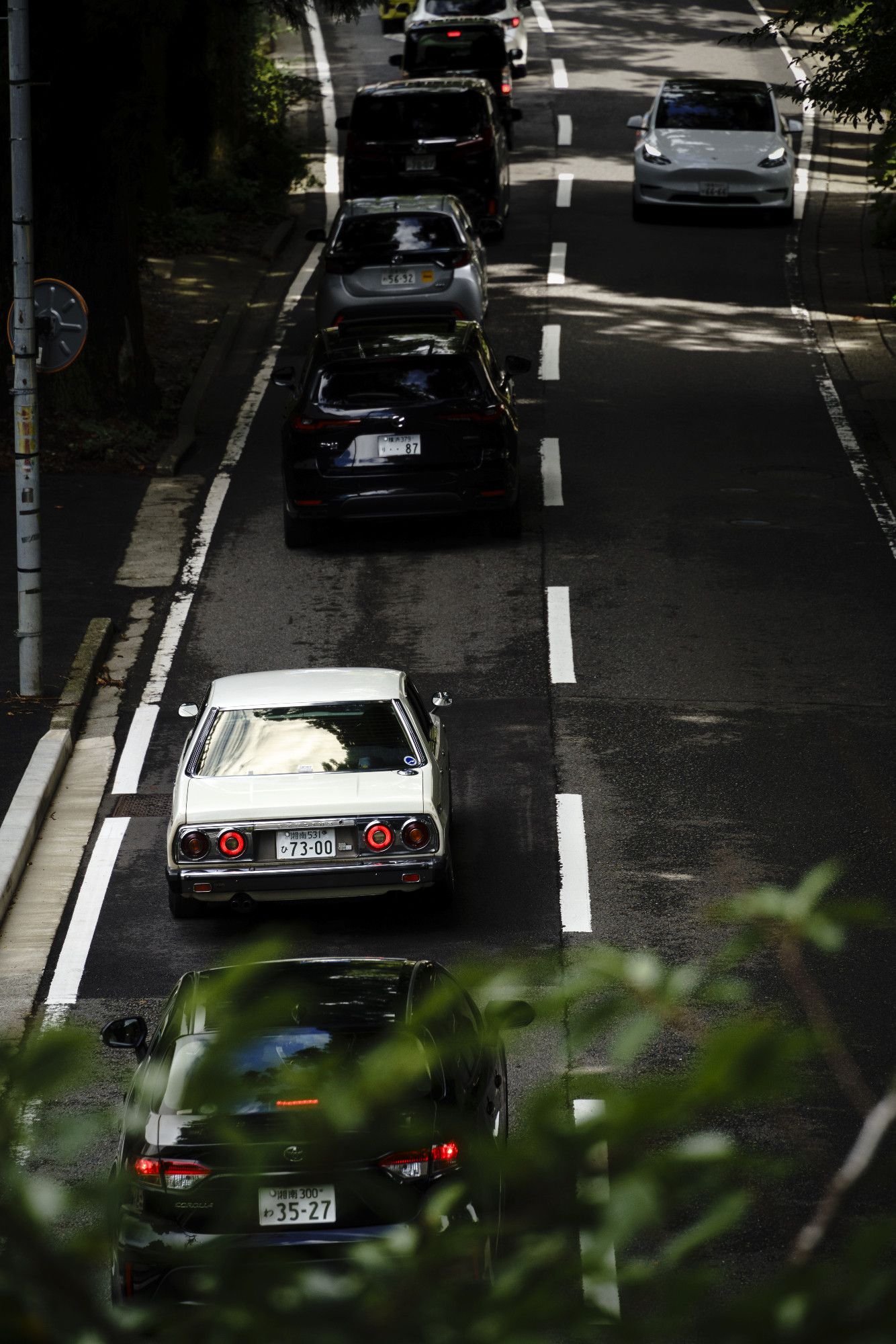 A white, classic Nissan Skyline sedan is on a Japan single two way road, in a traffic. A white Tesla is coming from the opposite lane. Under the Skyline there are green tree leaves blurred by the depth of field.