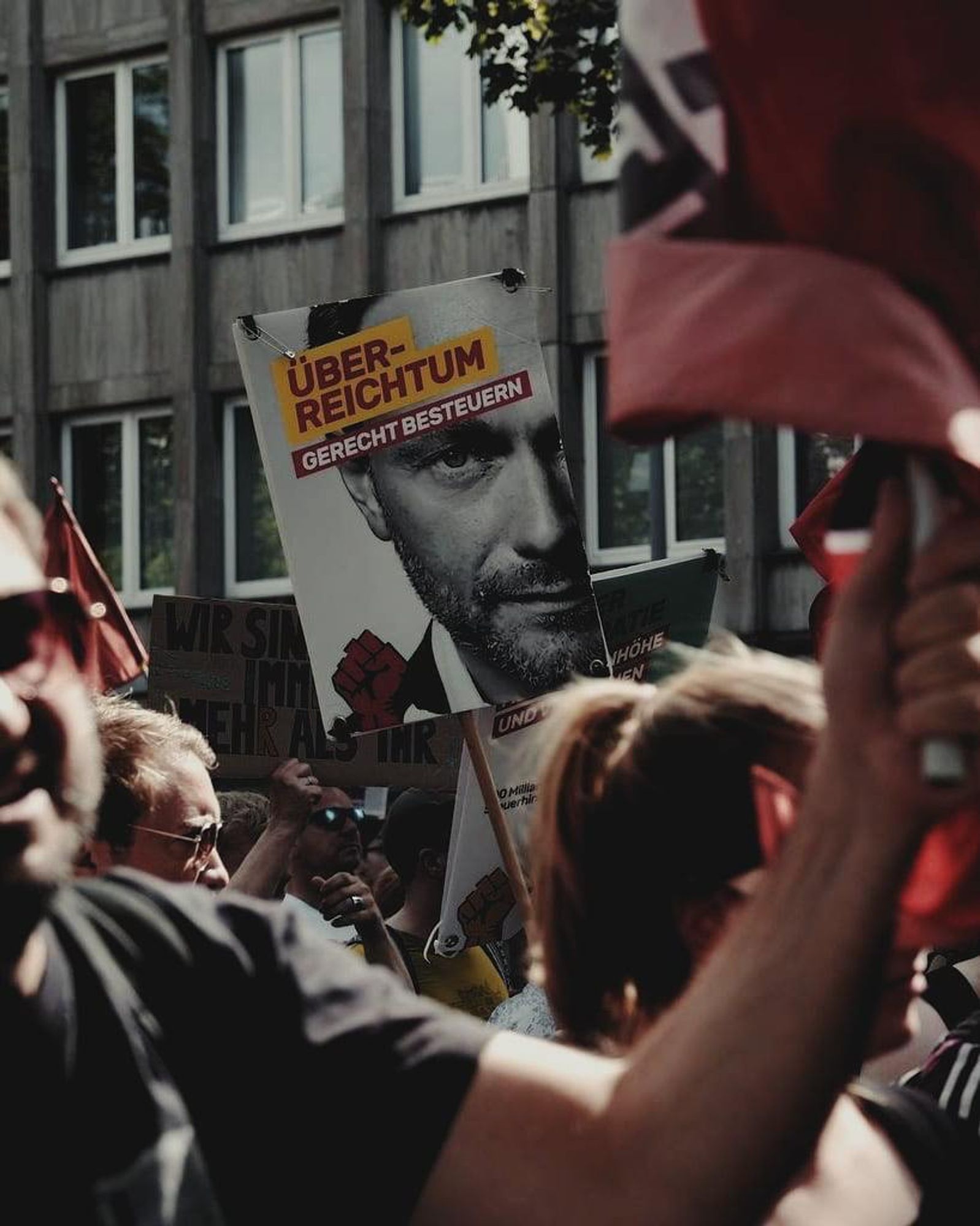 Foto: Demoimpression. Im Vordergrund werden friedlich Antifa-Flaggen geschwenkt. Im Fokus des Bildes befindet sich ein Protestplakat mit einem Schwarz-Weiß-Bild von Bundesfinanzminister Christian Lindner und der Forderung “Überreichtum gerecht besteuern”