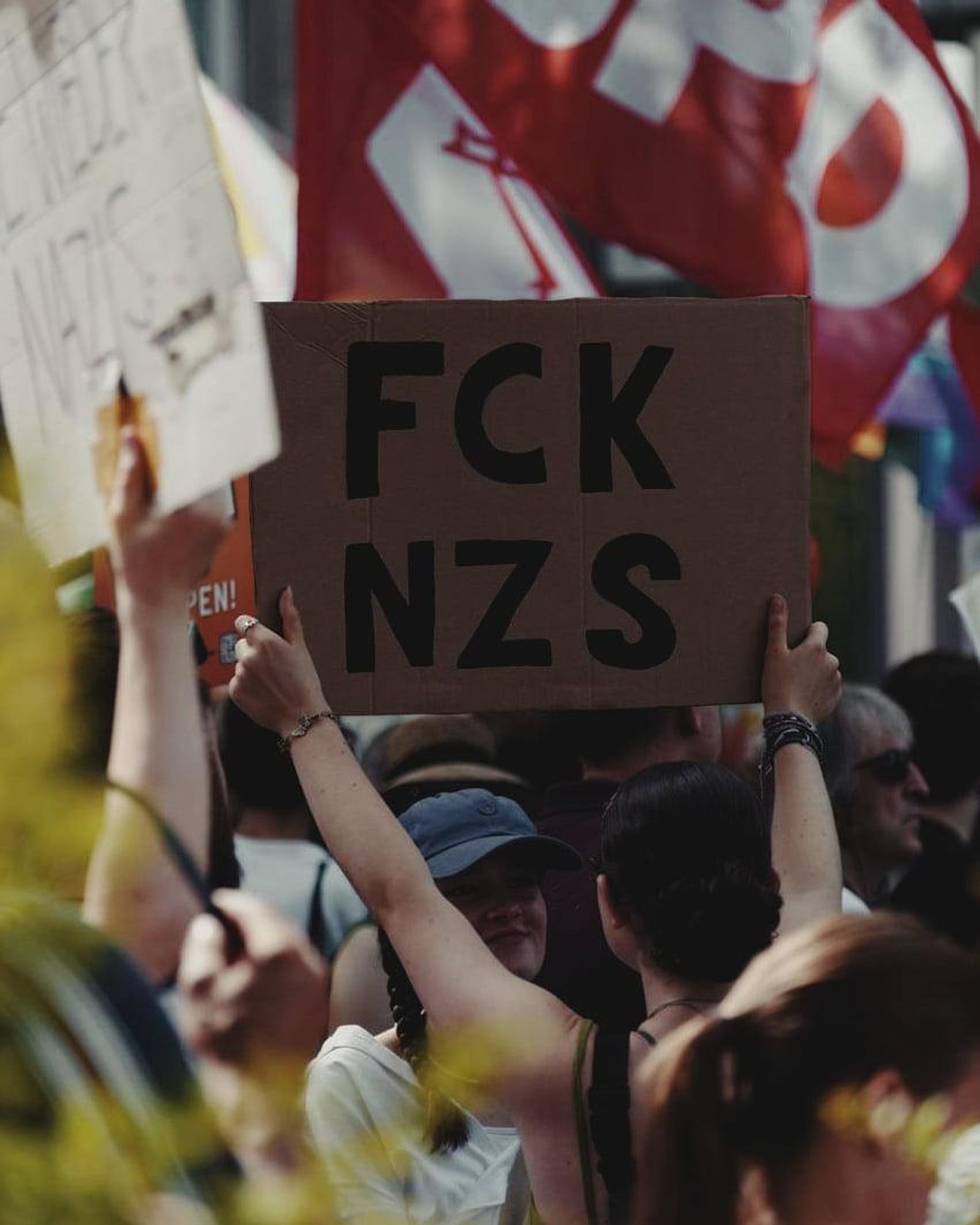 Foto: Demoimpression. Im Fokus befindet sich eine Frau, die ein Protestplakat mit dem Rücken zur Kamera in die Luft hält. Mit schwarzem Edding steht in fetten Buchstaben: FCK NZS (Fuck Nazis) geschrieben.