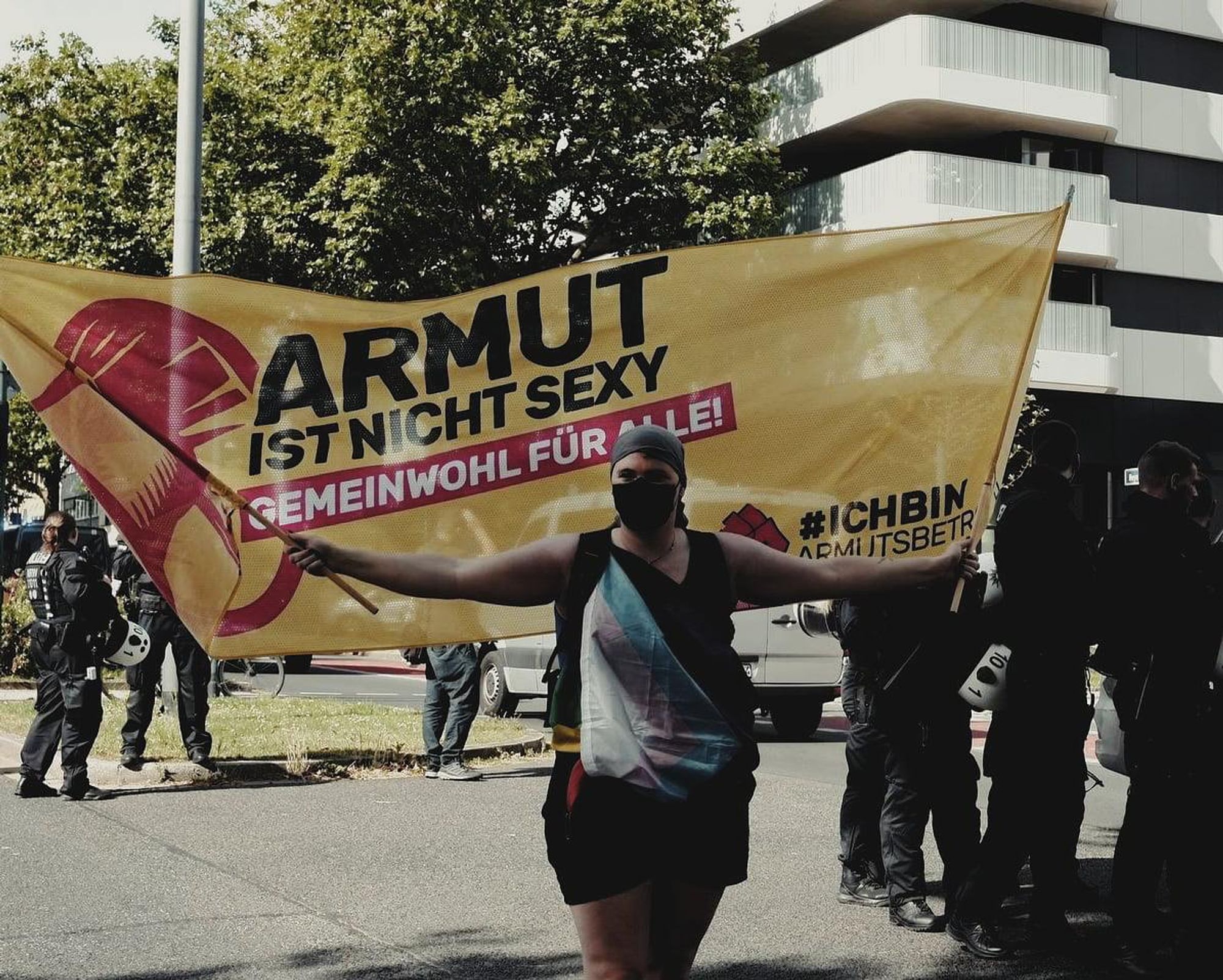 Foto: Aktivistin umwickelt mit der Prideflagge und schwarzer FFP2-Maske schwenkt friedlich ein gelber Protest-Banner. Ein rotes Megafon ist abgebildet mit dem Text: Armut ist nicht sexy - Gemeinwohl für alle! #IchBinArmutsbetroffen und einer roten Protestfaust. Im urbanen Hintergrund stehen Massen an uniformierten Polizeibeamten:innen.