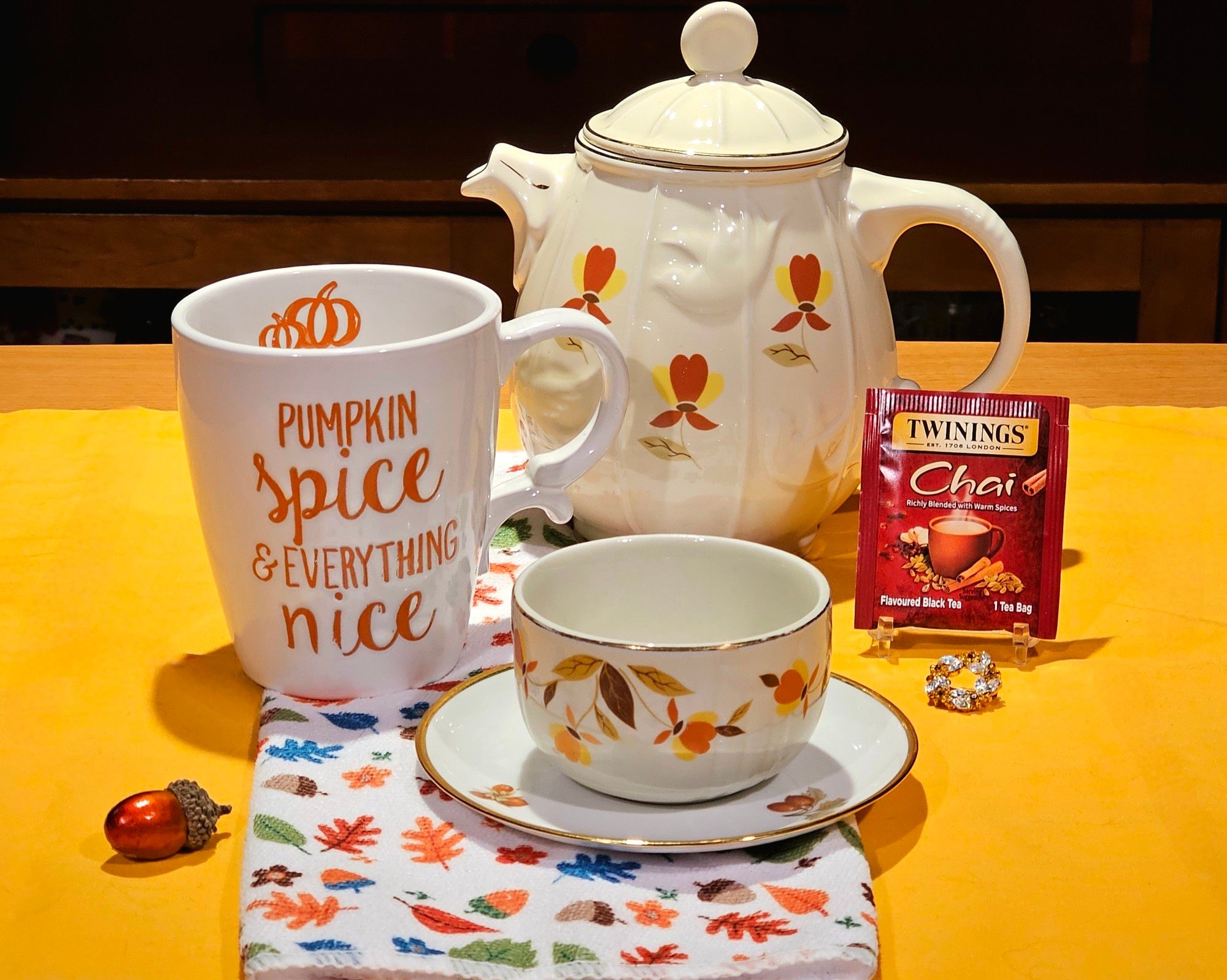 A tea tableau in comforting warm colors of saffron, cinnamon, cream, pumpkin, copper, and teal. An heirloom teapot & bowl with falling leaf patterns accompany a mug that says "Pumpkin Spice and Everything Nice" and there is a shiny acorn and a small, jeweled citrine & blue topaz wreath brooch, all on a tea towel with a cheerdul pattern of leaves & acorns in Fall colors. All of this sits on a bright saffron yellow cloth. The tea is Chai Spice black tea from Twinings.