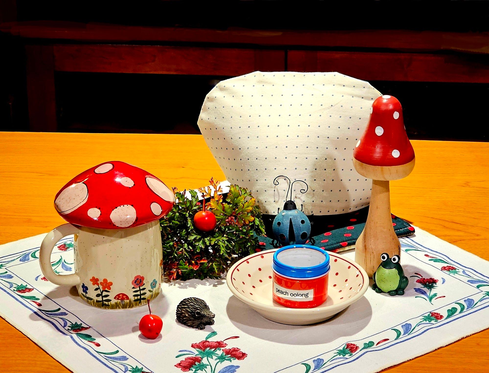 A tea tableau with dotted things, mostly white with red, such as a polka dotted bowl, a mug shaped like a toadstool, & a wooden toadstool, and a dotted tea cozy on a dotted hot pad, surrounded by a blue ladybug, a goofy green frog, a wee hedgehog, two tiny crab apples, and a cluster of Herb Robert, all on a tea towel with a red, blue, & green vine border. The tea is Peach Oolong in a red & bright blue tin.