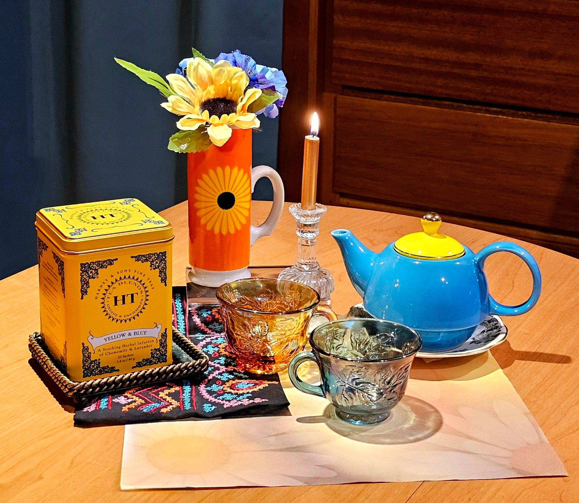 A tea tableau tribute showing hope and light for Ukraine. On a mat designed with sunflower silhouettes there is a square of traditional embroidered cloth, and china & glassware in yellows & blues to commemorate the Ukrainian flag, as well as a tall cup with a sunflower image on it and a sunflower and spray of blue flowers inside it, and a lit golden candle on a crystal candlestick. The tea is called Blue And Yellow, from Harney and Sons, and a dollar from every purchase goes to support Ukrainian relief. Slava Ukraini.