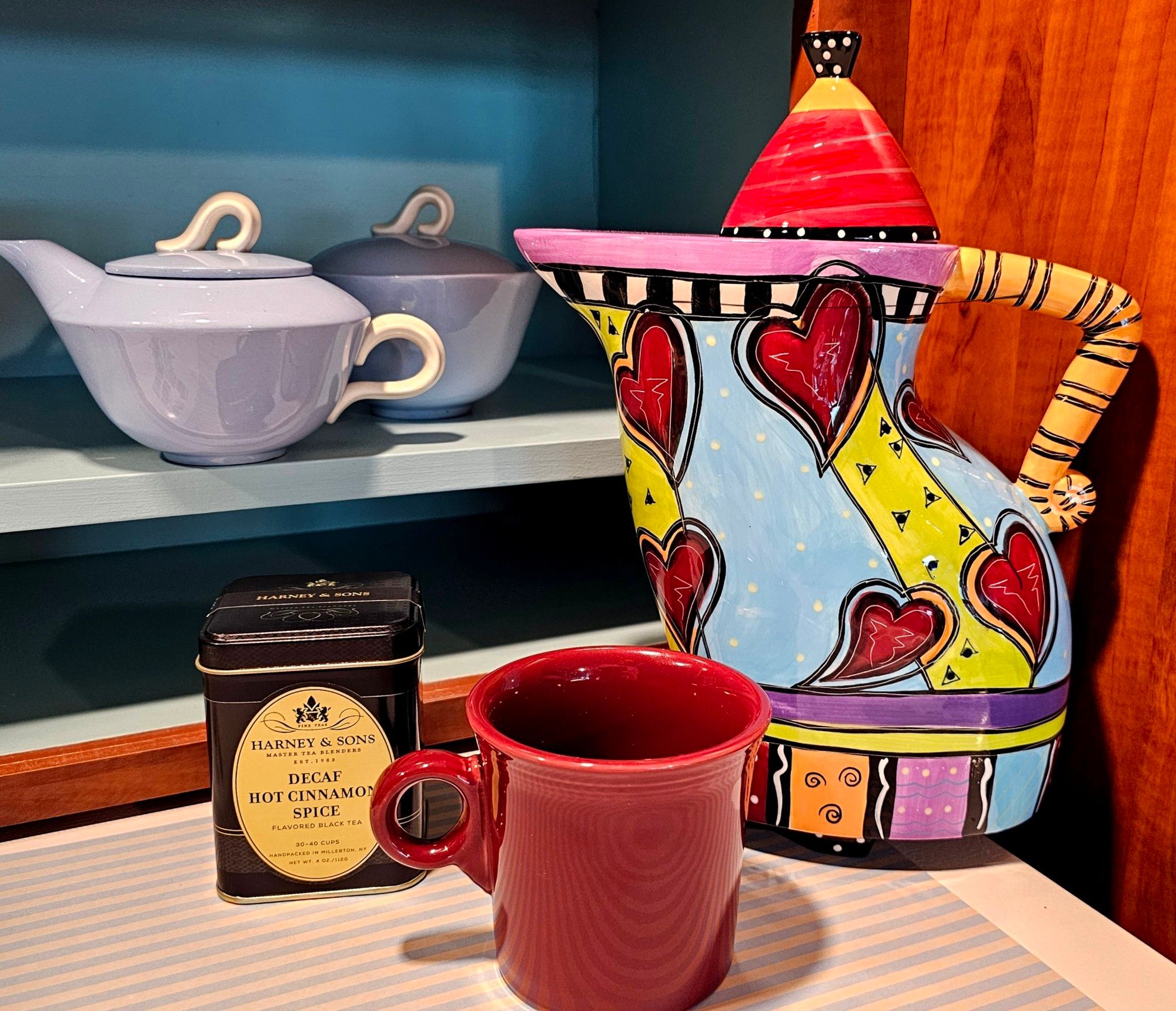 A tea tableau with a very tall coffee/tea pot in a crazy quilt of colors and lines. The pot is angular and quirky. There is also a maroon Fiestaware mug and a pale blue creamer & sugar bowl that look like Aladdin's lamp. The tea is Decaf Hot Cinnamon Spice from Harney and Sons.