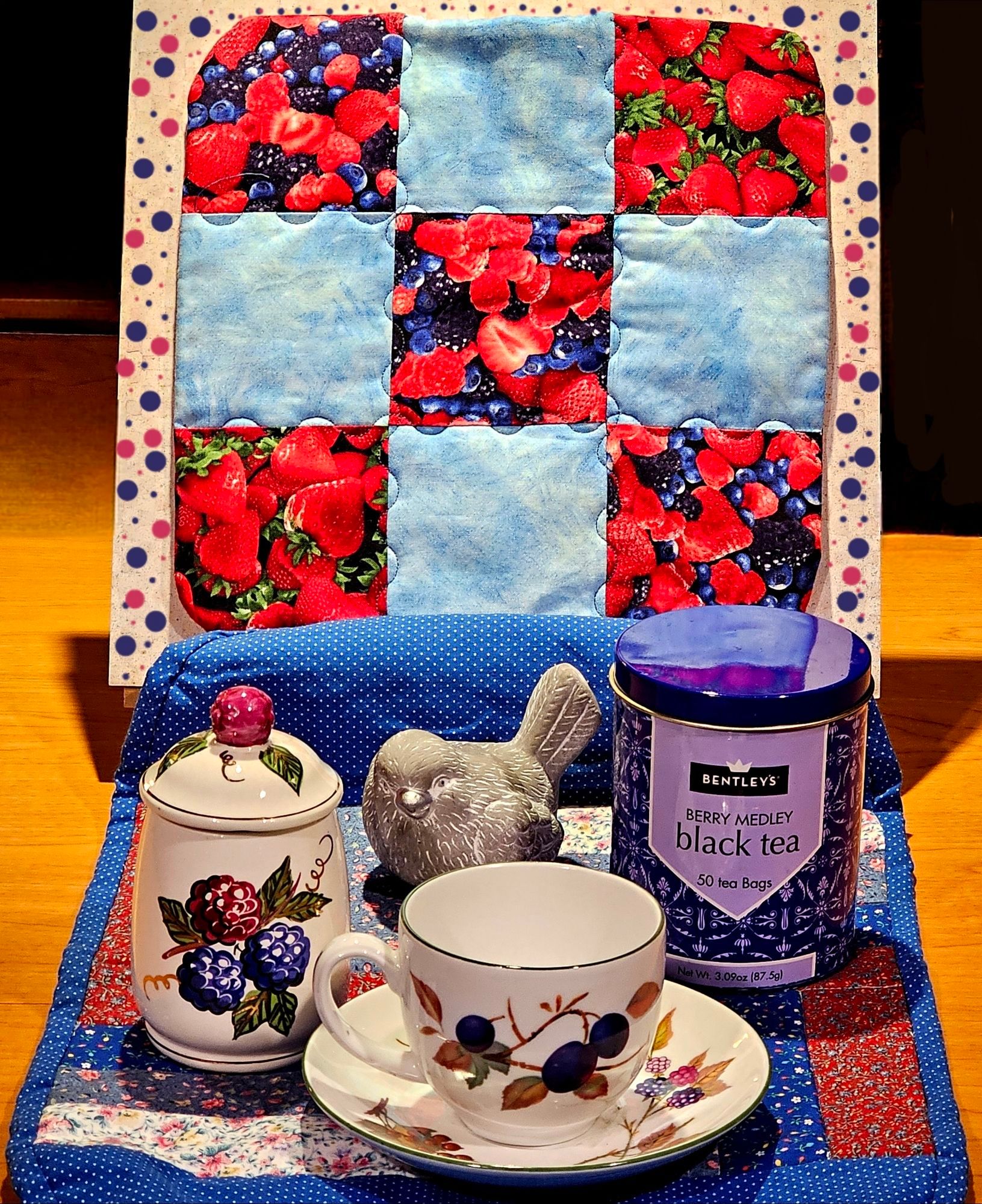 A tea tableau in deep blues & reds with a berry theme, including berry designs on quilt squares and china. There is a carved stone bird overseeing everything, and the tea selection is Berry Medley black tea from Bentleys.