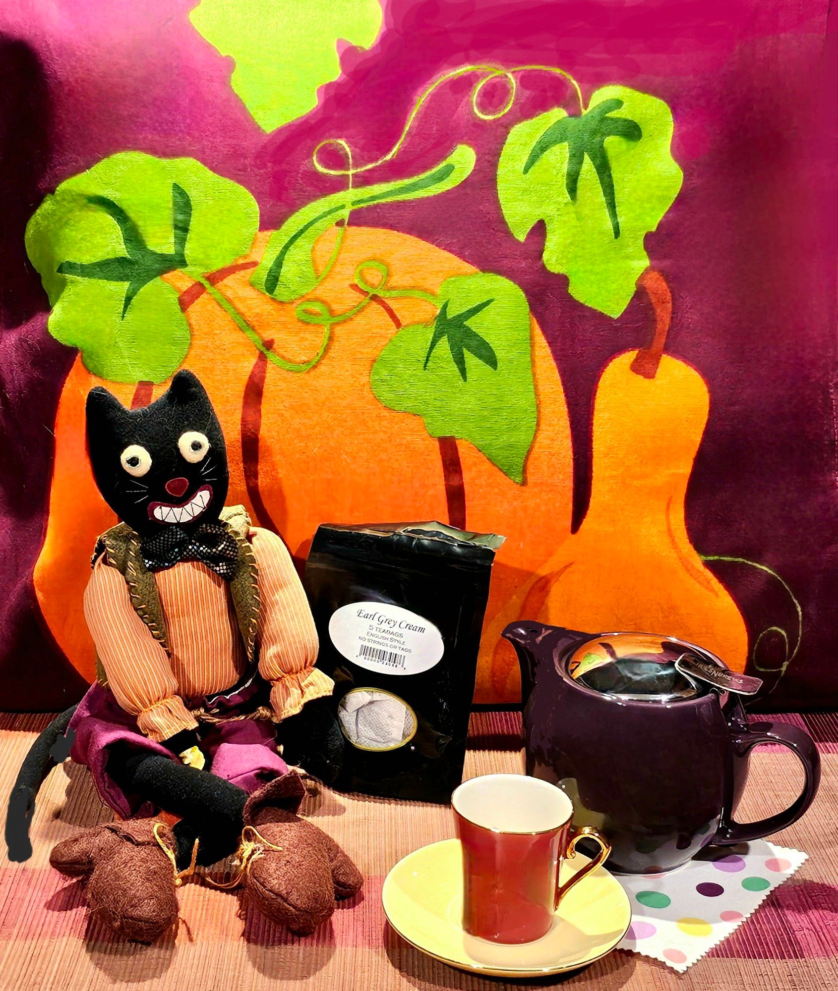 A tea tableau in bright Fall colors balanced by black & deep purple. Against a fabric backdrop depicting pumpkins & squash we have a purple teapot, a vintage demitasse set, and a hand-sewn felt black cat in rustic peasant clothing and big boots, with a large, toothy grin on its face and round, staring eyes. The tea is Earl Grey Cream, which makes a nice London Fog with the addition of lavender syrup & steamed milk foam.