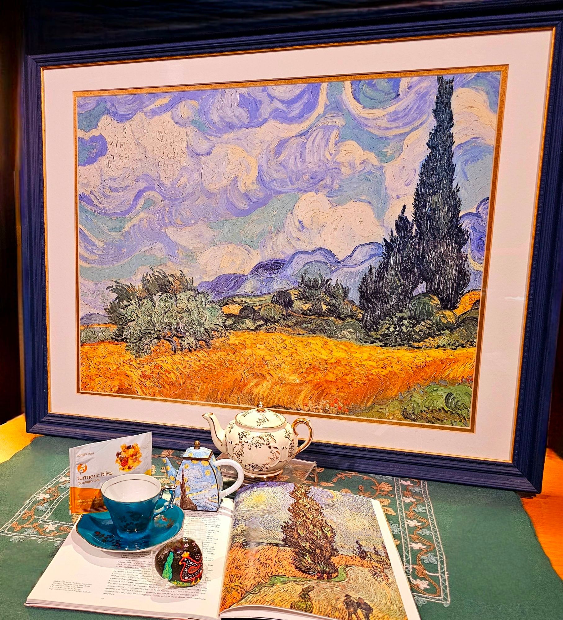 A tea tableau with a cypress tree theme featuring works by Van Gogh, and a painted rock with a cypress tree & a folk art chicken. The tea is Turmeric Bliss herbal from Adagio.