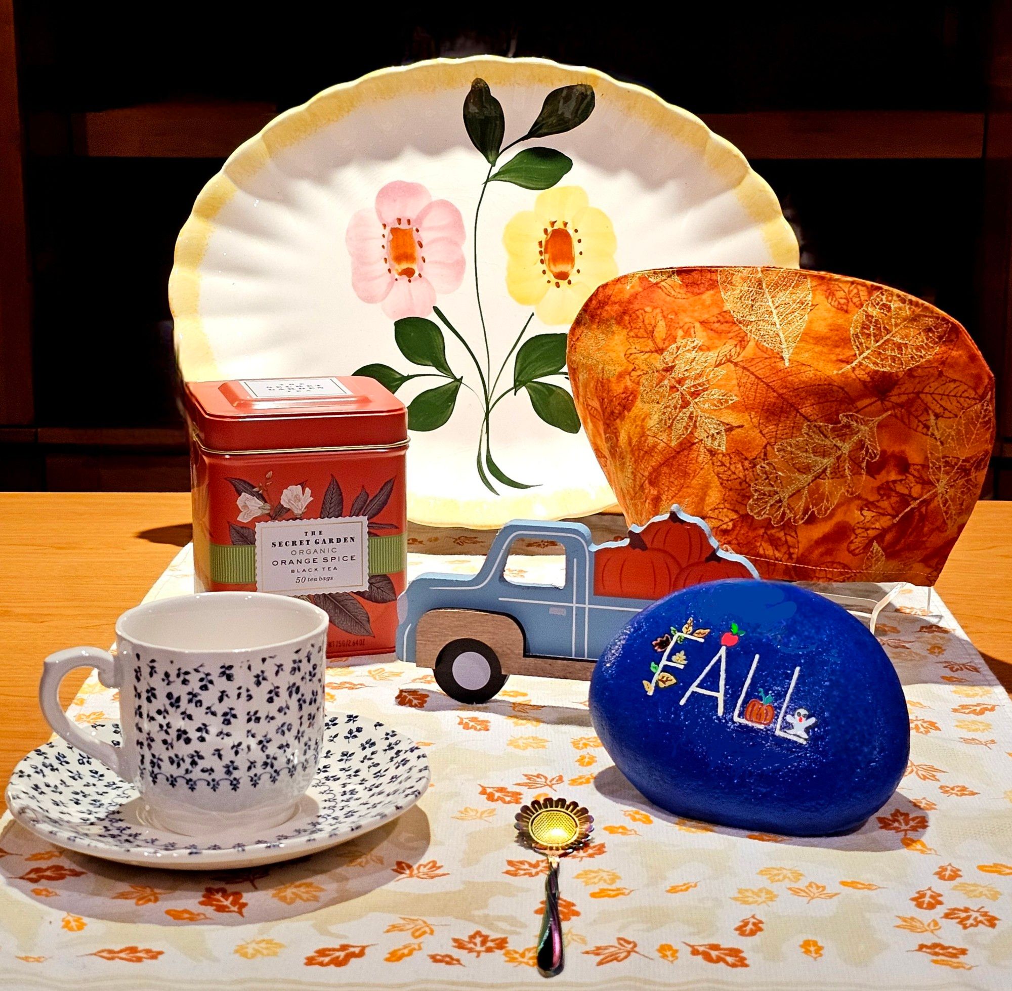 A light-filled tea tableau with crisp points of Fall color and touches of blue, like a day in early Autumn. There is a wooden truck carting pumpkins, a rock painted with the word "Fall" where each letter is occupied by leaves, an apple, a pumpkin, or a scary little ghost. Vintage china, a sunflower spoon, leaf-patterned linens, and an orange tin of Orange Spice tea complete the display.