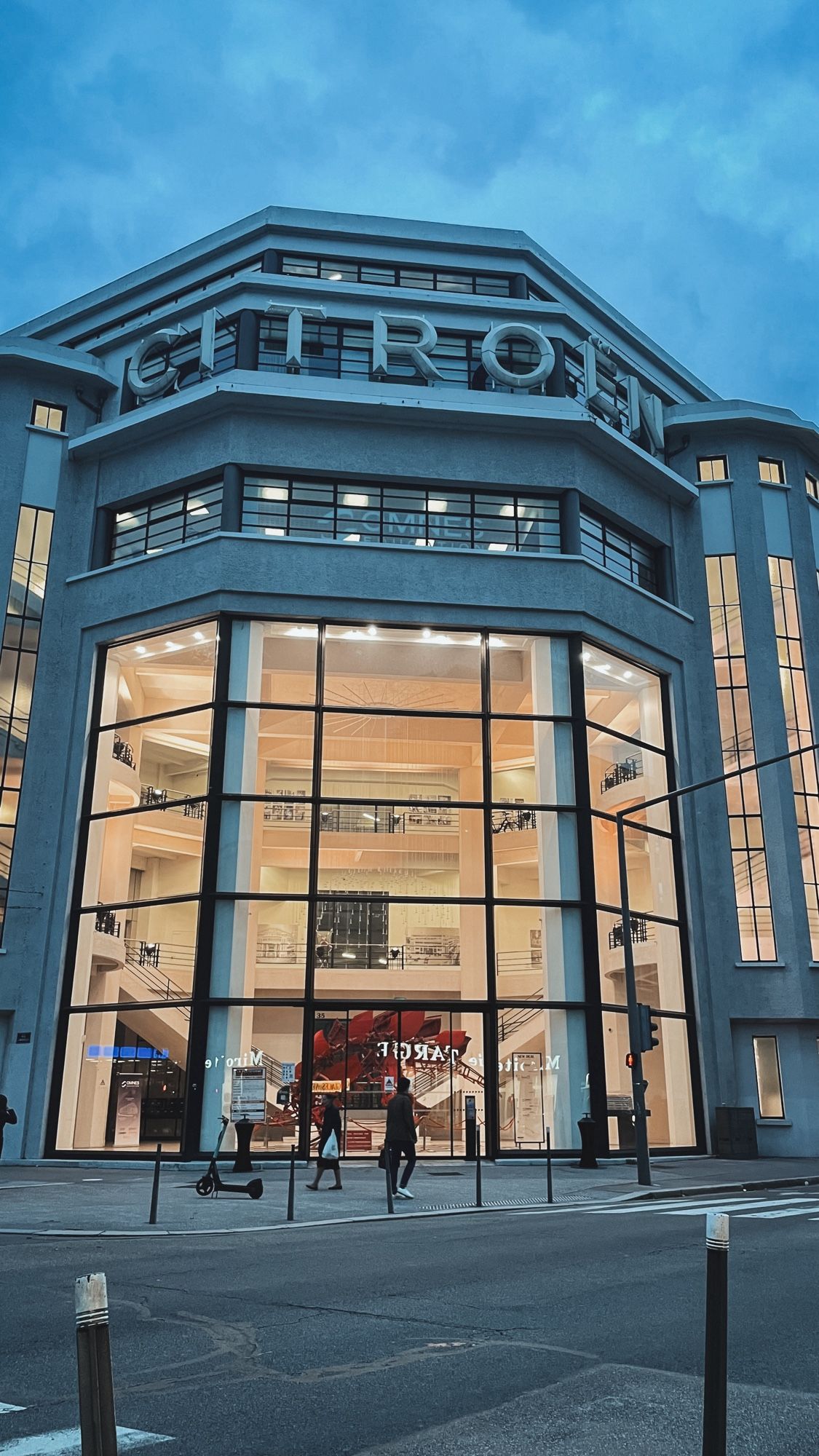 Un bâtiment pris en photo à la tombée de la nuit. Ils est constitué de plusieurs étage et une immense « vitrine » permet de voir l’intérieur. En haut, en lettres immenses on voit écrit « Citroën »