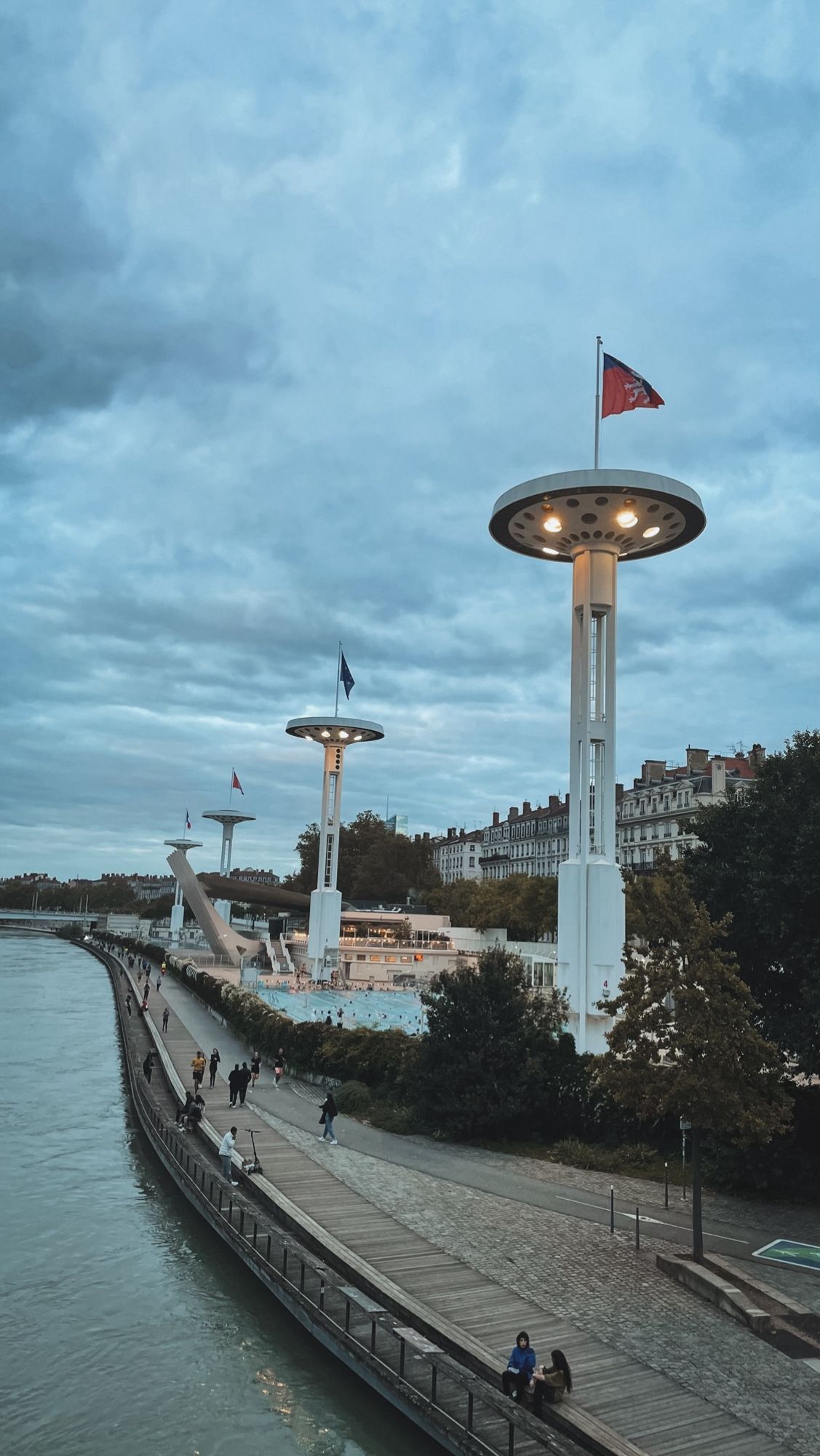 En bord du Rhône on voit une promenade. Des gens se promènent, au second plan dans la perspective on voit le stade nautique en bord du Rhône, avec sa piscine extérieure