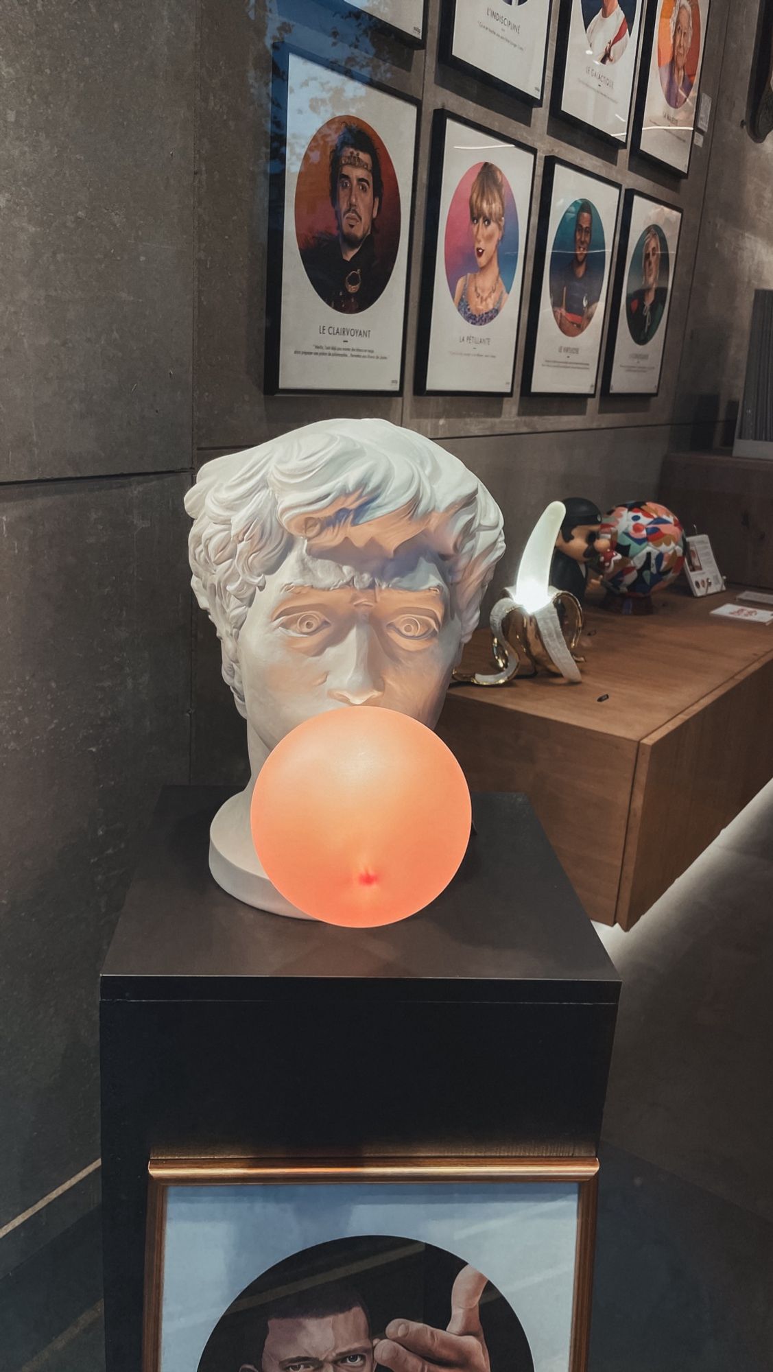 Photo d’une création artistique dans un boutique. 
Il s’agit d’une sculpture de tête d’homme faisant penser à Apollon. Il a l’air de faire une grosse bulle avec son chewing-gum et c’est en fait une ampoule qu’il tient dans sa bouche.