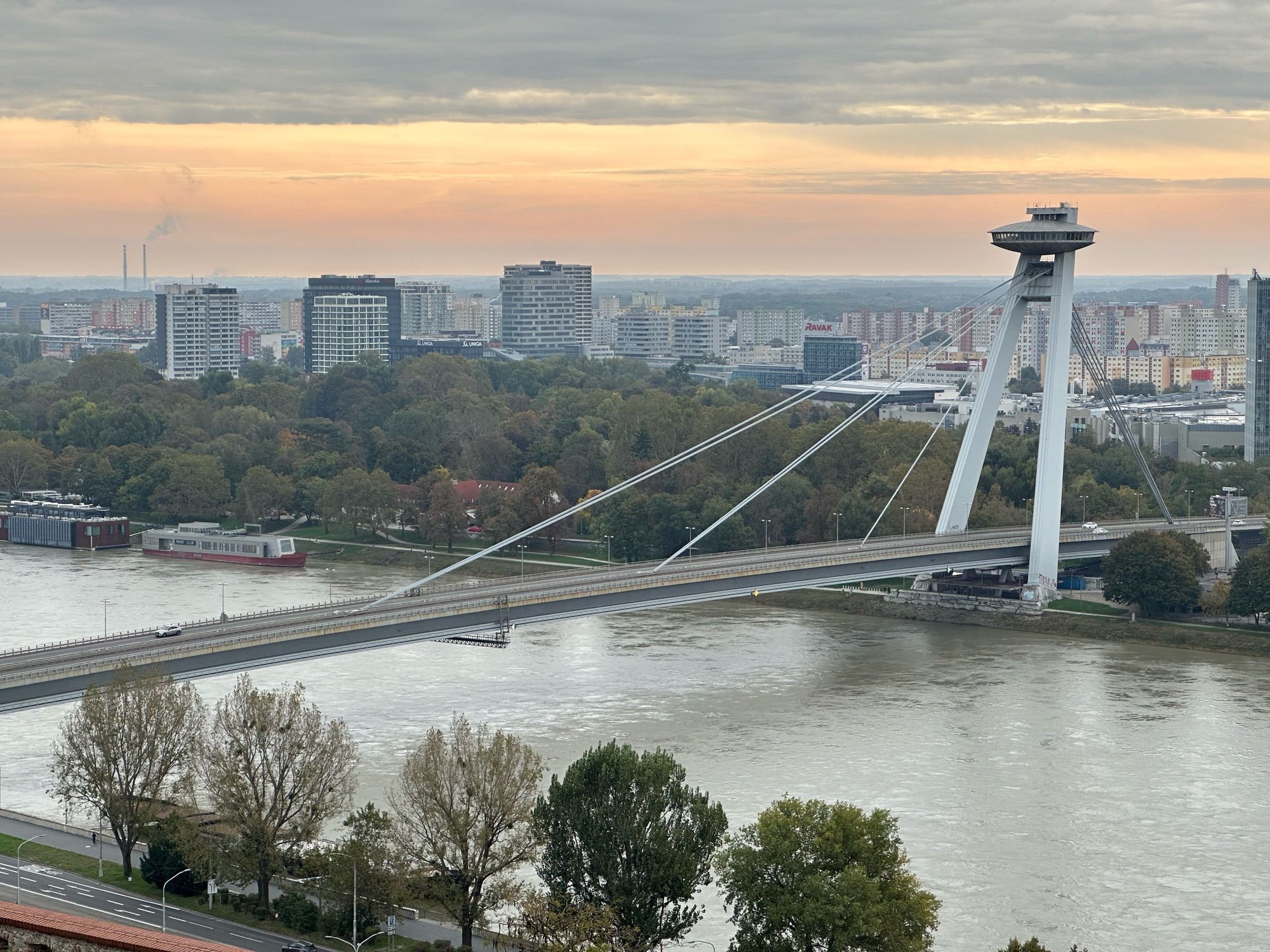 The Most SNP bridge as seen from Bratislava Castle. The sky is orangey-yellow in colour. Communal residential buildings are in abundance in the background 