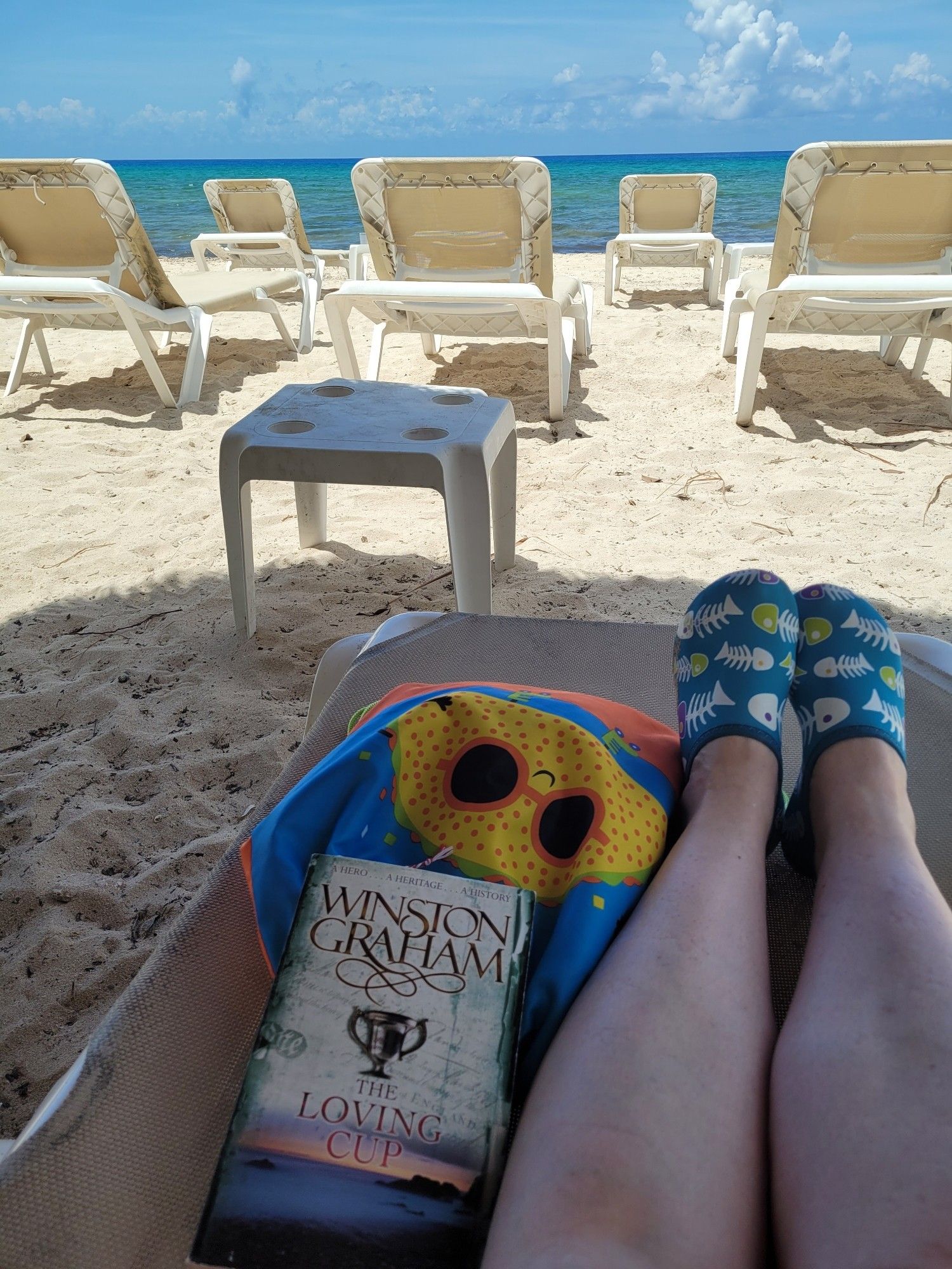 A shot looking out over a line of beach chaises, with the ocean on the horizon. In the foreground, Winston Graham's The Loving Cup rests on a beach bag with a happy taco design. Two feet in colorful fishbone printed water shoes are resting next to the bag.