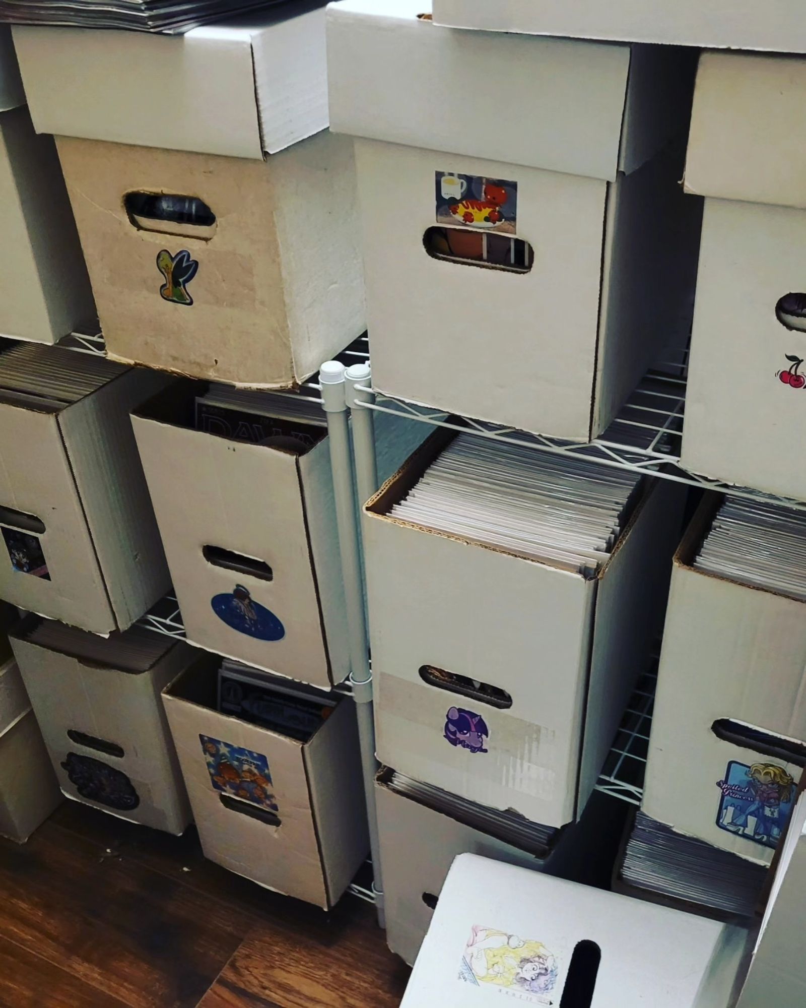 Many white cardboard short boxes full of comics on a set of short white wire racks. These boxes also all have a cute sticker on the front of each box. (To know which is the front end!)