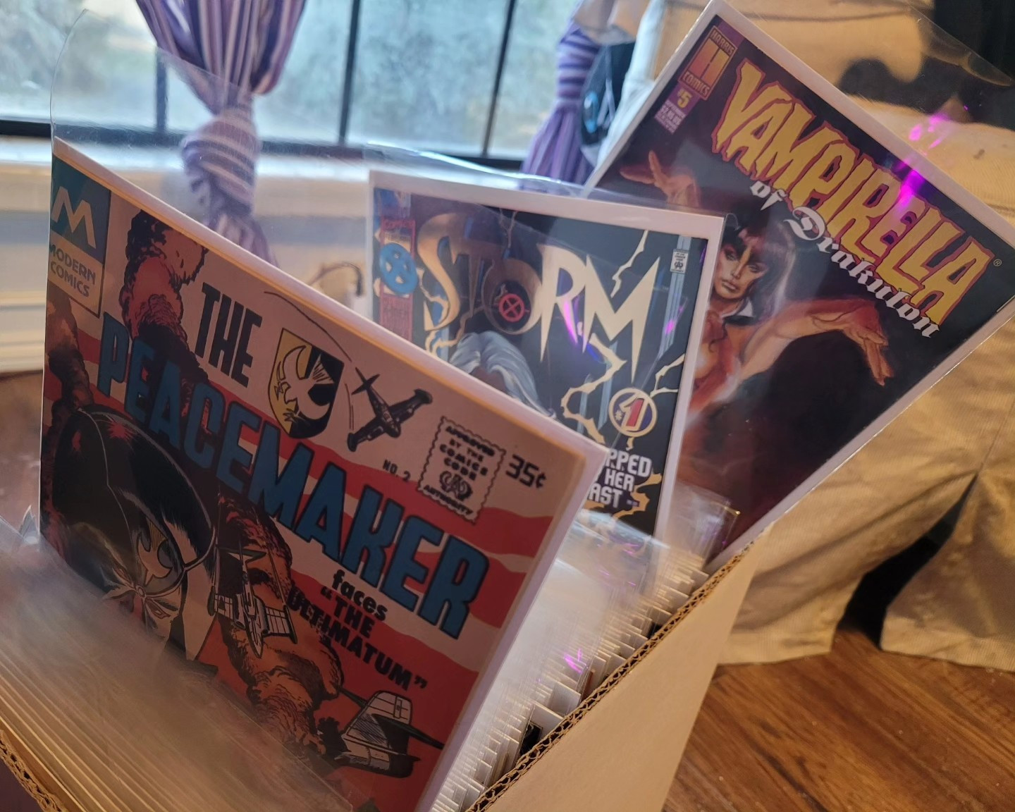 A plain white short comic box full of bagged & boarded comics. The tops of a Peacemaker comic, Storm comic and Vampirella comic are poking up over the edge of the box.