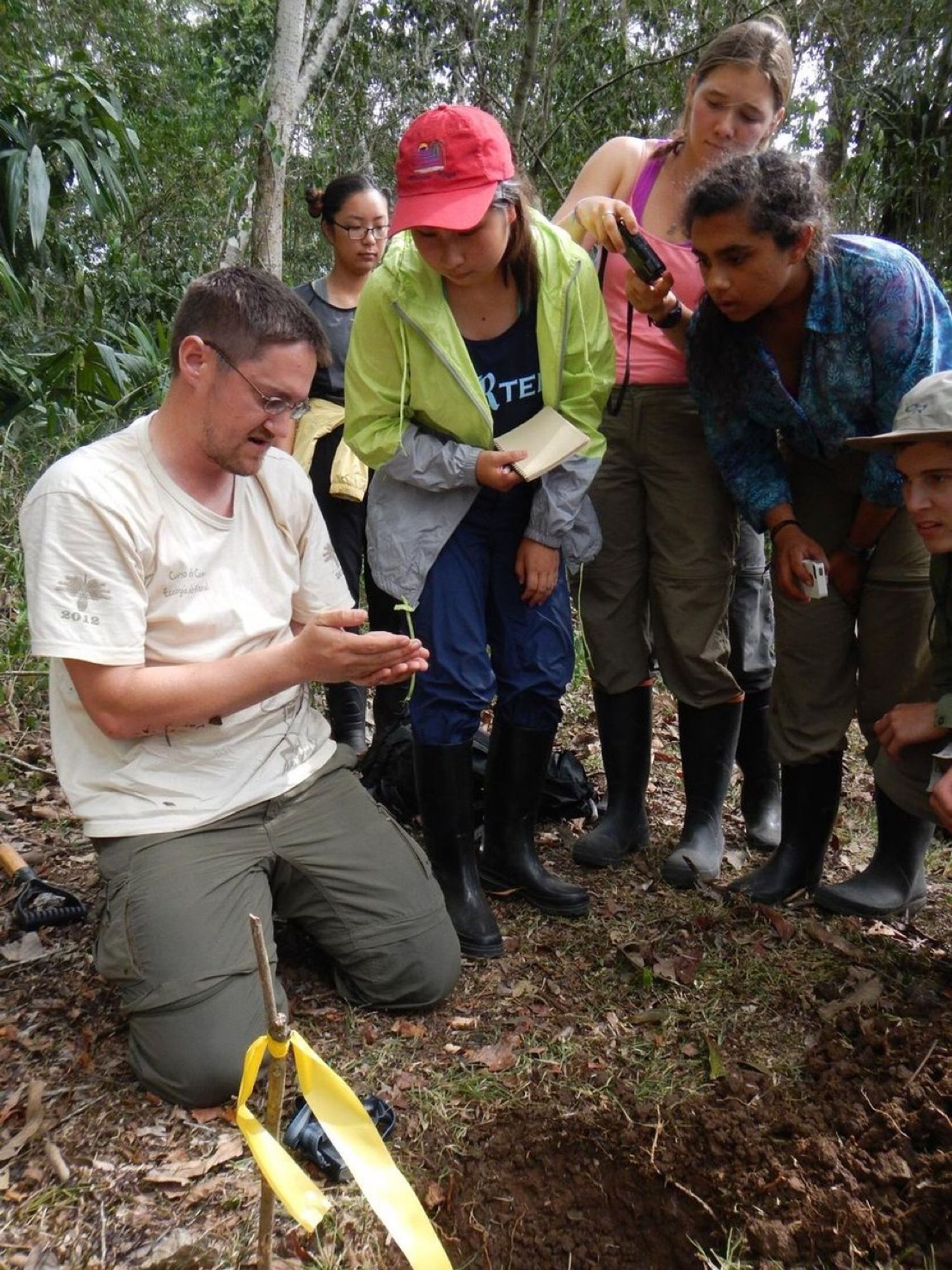 A field biologist on his knees in the rainforest showing something in his hands to a group of eager students