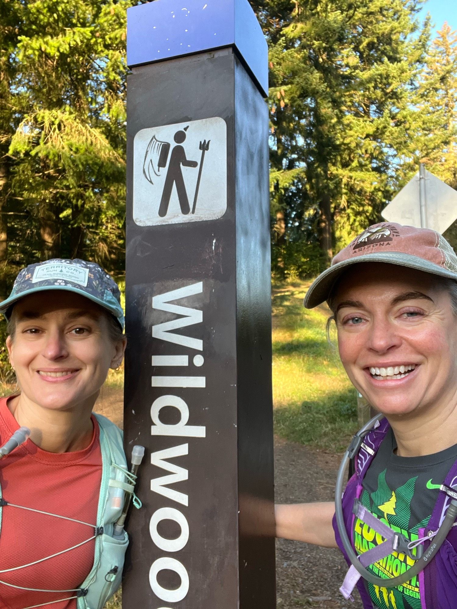 Two white, middle aged women taking a photo at the start of the Wildwood trail near the zoo/arboretum in Portland, Oregon with a sign saying Wildwood in the background.