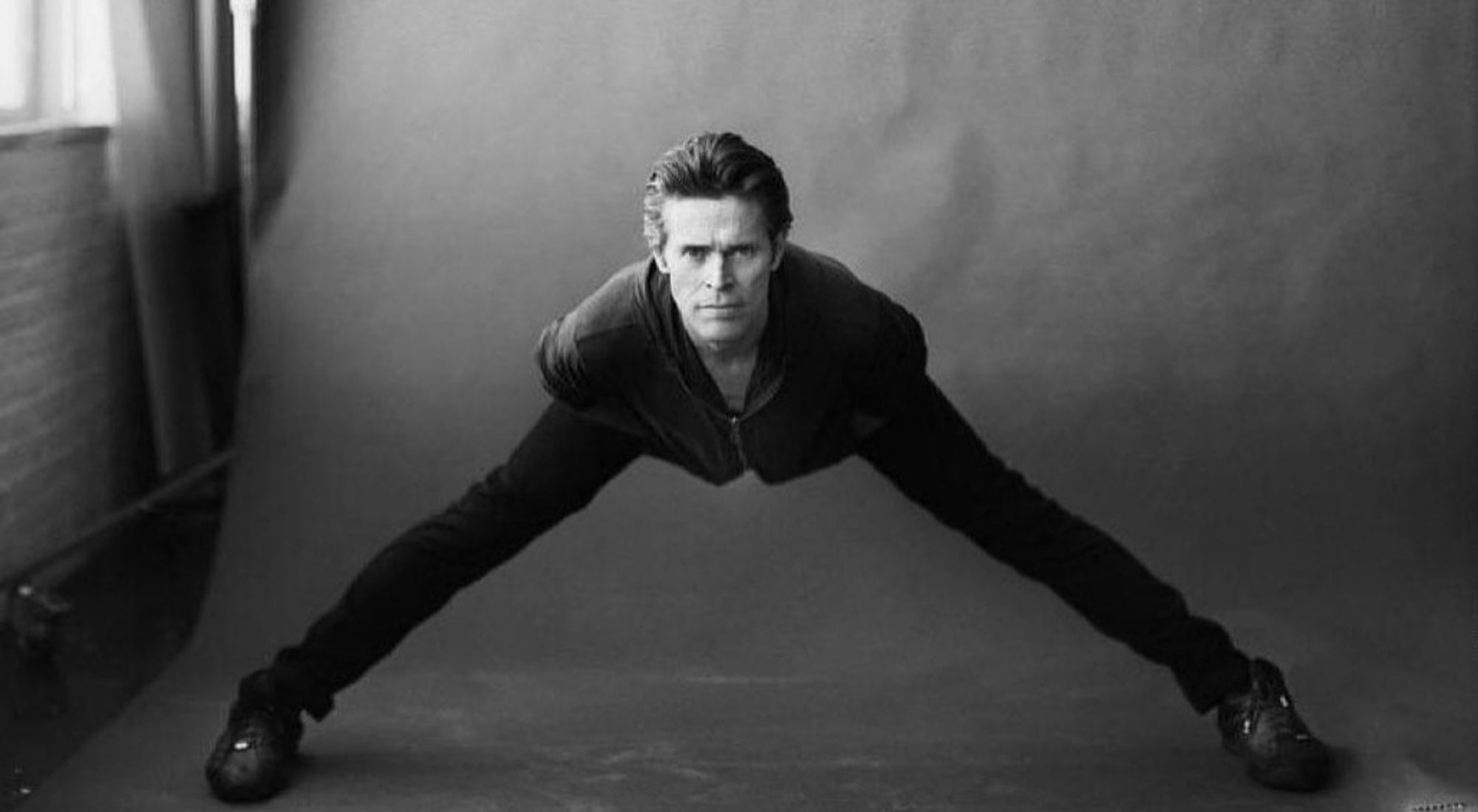 Willem Dafoe with his legs spread uncomfortably far apart, his chest tilted forward, and his face toward the camera