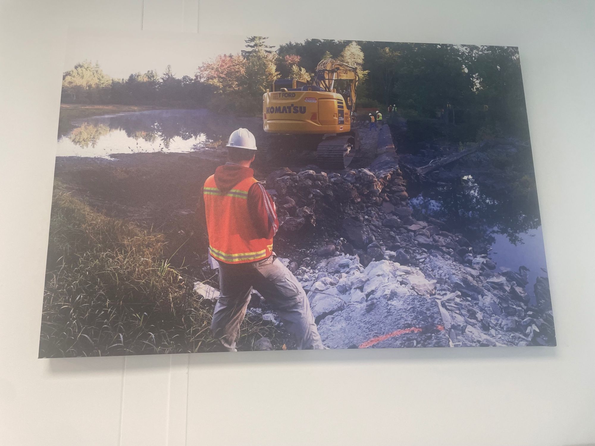 Restoration professional in high-viz vest and hardhat observing a dam removal construction site containing partially demolished dam, construction workers and a large excavator.