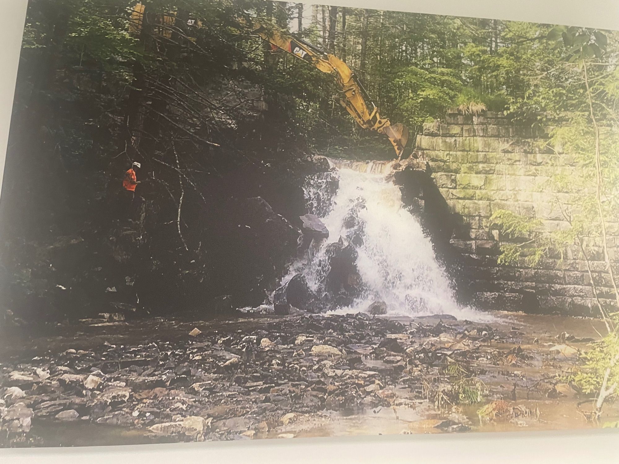 A large excavator in the process of notching a dam, a slow method in which an impoundment is drained through notches cut into the dam. New notches are cut in so the water drains out of the reservoir at a consistent flow. The sediment trapped behind the dam flows downstream in a fixed rate that allows the ecosystem to adjust to the changes.