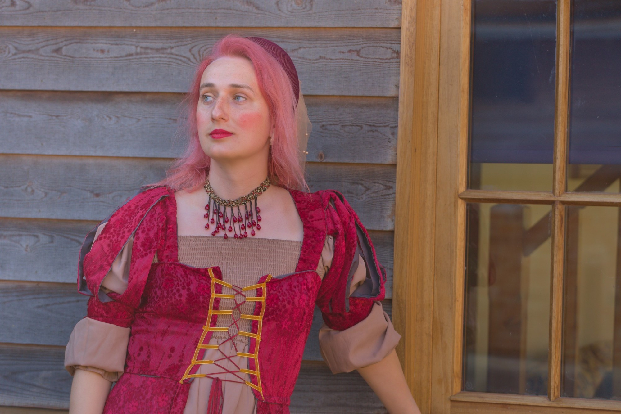 Heather, a pink haired woman stands in front of a wooden wall, looking off into the distance. She wears a red slashed dress with a slight dripping pattern, laced at the front, over a plain pale brown dresss. Around her neck is a choker choker styled to look like dripping blood.