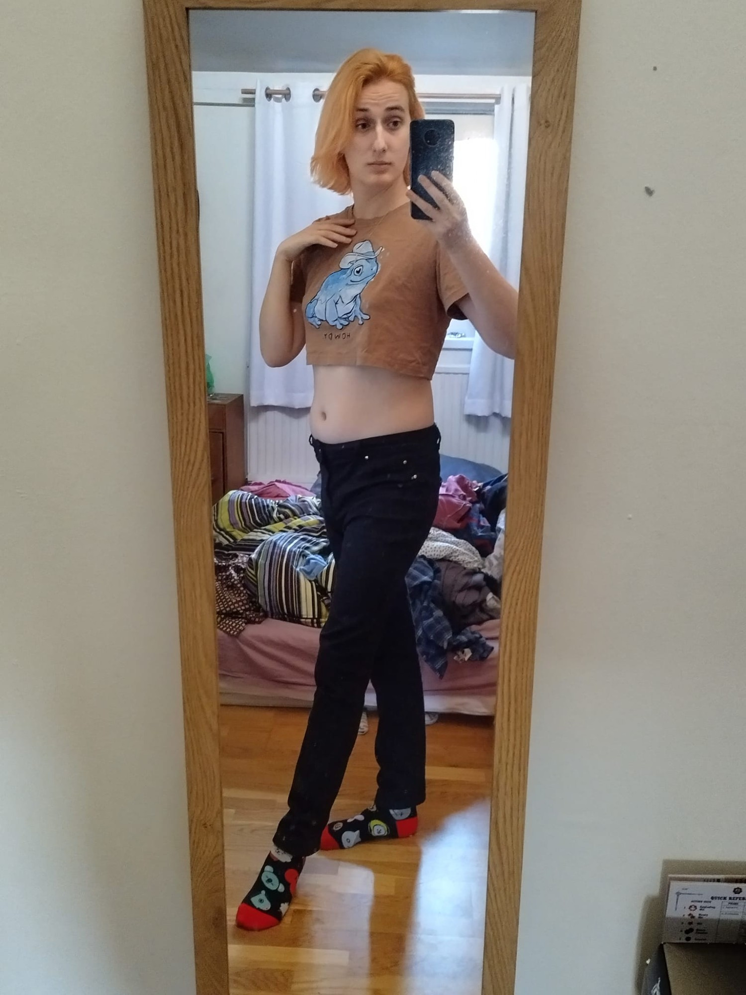 Heather, a white woman with peach colour hair, stands in front of a mirror. She is wearing black skinny jeans, and a brown crop top with a blue frog in a cowboy hat on it. Her room is disgracefully messy behind her.