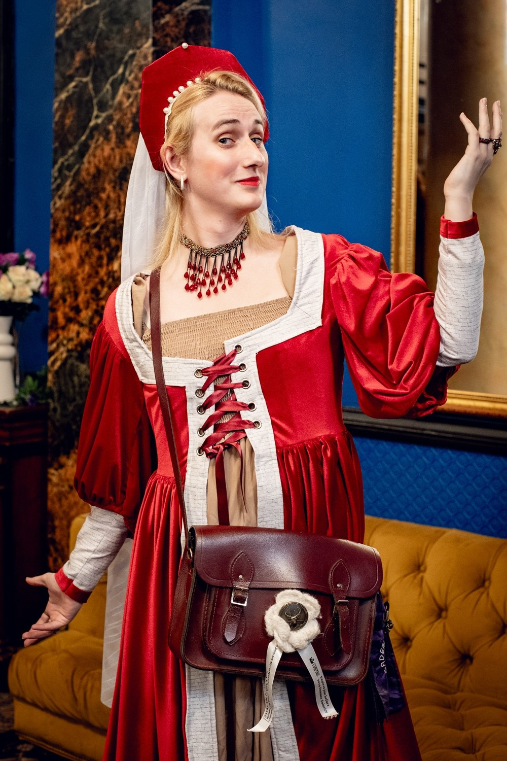 Heather, a white woman, poses for the camera smugly, in front of a blue wall and stone pillar. She is wearing a red velvet dress, in a tudor cut, with white trim featuring a brickwork motif. She wears an angular hat, with pearls, from which drapes a long white veil.