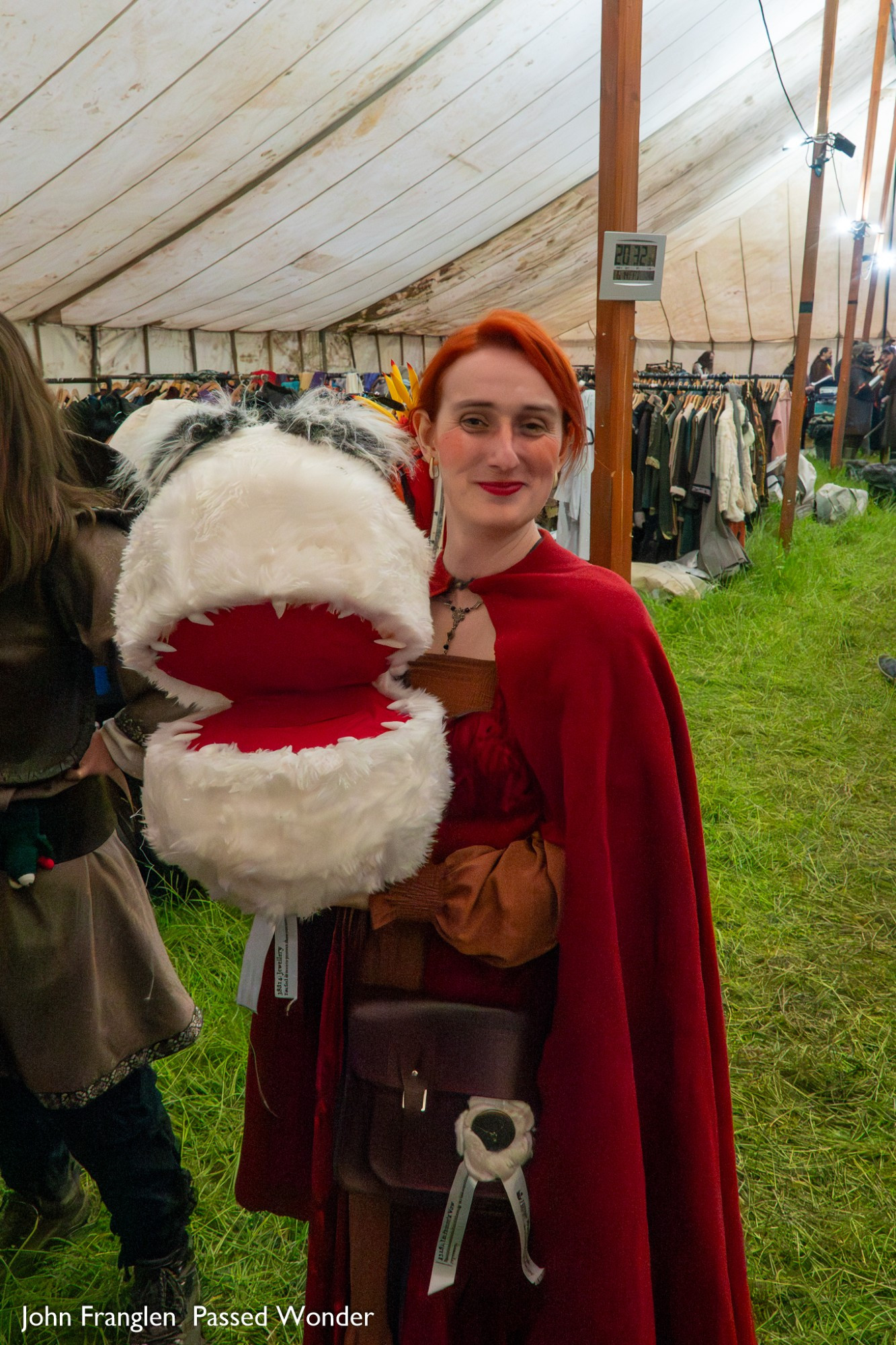 Heather, a red haired woman, stands in a tent filled with costume racks. She wears a red cloak, over a flashed red dress. In he hands is a large round puppet, roughly 2 feet long. It is white and fluffy, with massive fluffy eyebrows. Its mouth is open and sharp teeth are visible inside