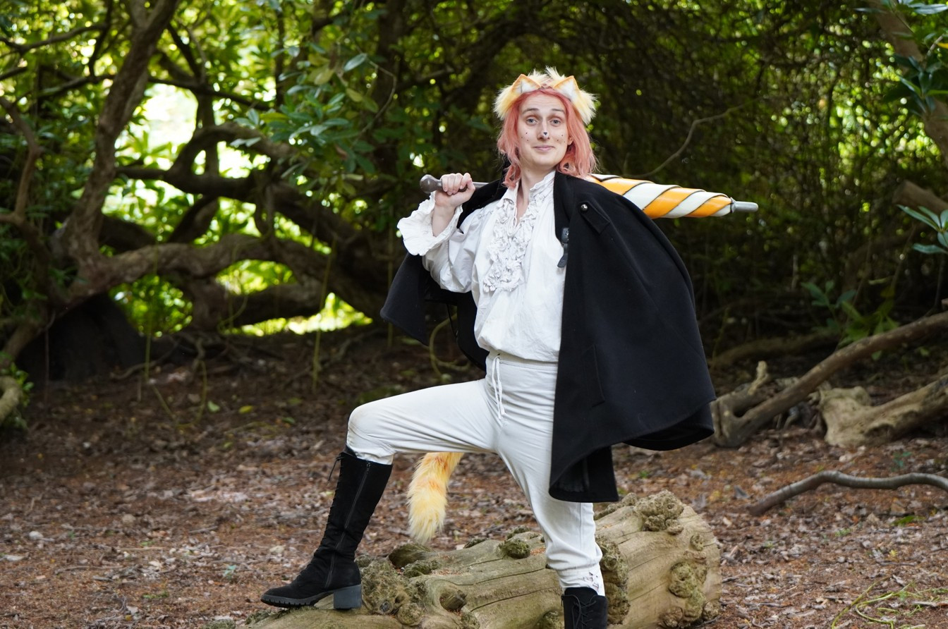 Heather, a pink haired woman is dressed as Puss in Boots standing dramatically in a wood, one leg raised on a fallen tree. She has orange and white cat ears and a tail, and is holding an umbrella in the same shades. She is wearing a white frilly shirt and a pair of tight trousers. Around her shoulders is a heavy black cape. She looks down the camera and smiles a cheeky grin.