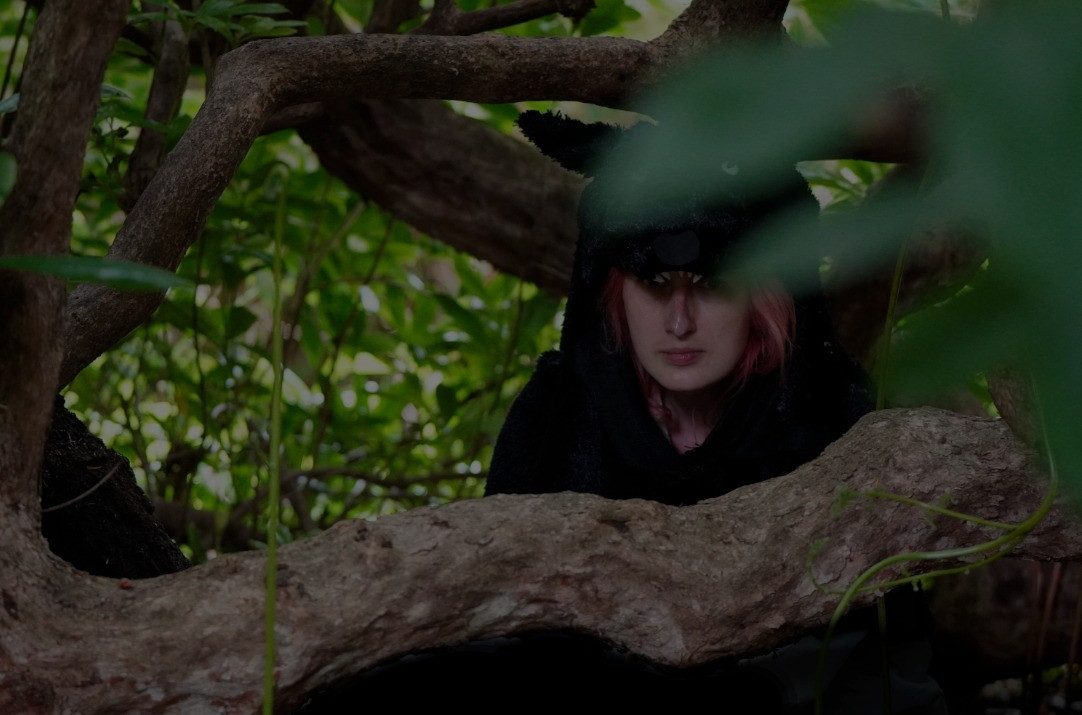 Heather, a pink haired woman in deep shadow waits in a bush. Atop her head is a hard to see wolf mantel. Most of the costume is obscured by leaves and branches.