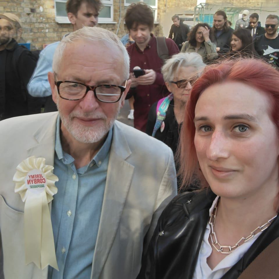 Heather, a red haired woman in her 30s takes a selfie with Jeremy Corbyn. She is wearing a leather jacket, white crop top and a chain. He is in a white suit, blue shirt and a vote Corbyn Rossette.