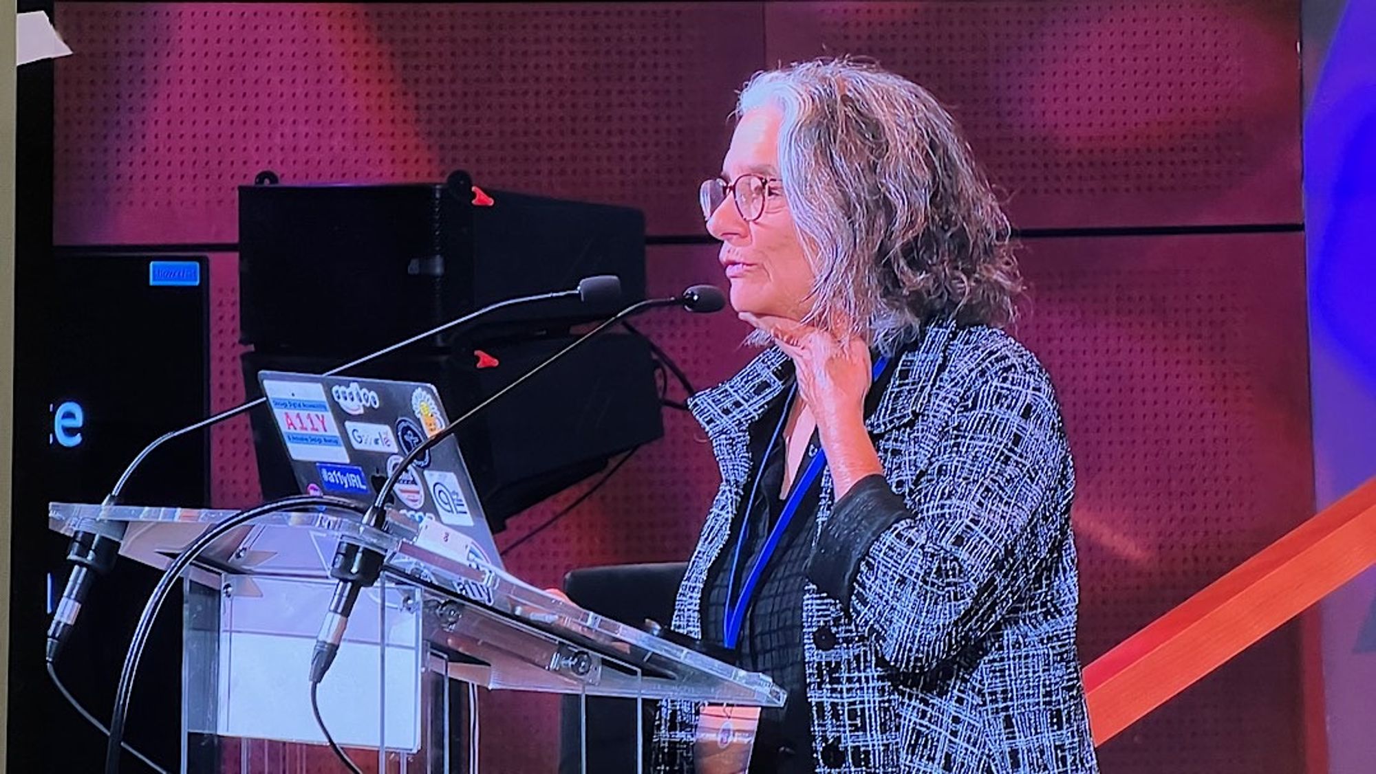 Un orateur fait une présentation sur un podium avec plusieurs microphones. Elle a les cheveux gris et bouclés et porte des lunettes, une chemise noire et une veste à carreaux. Un ordinateur portable avec divers autocollants est sur le podium. L'arrière-plan comporte un panneau rouge.