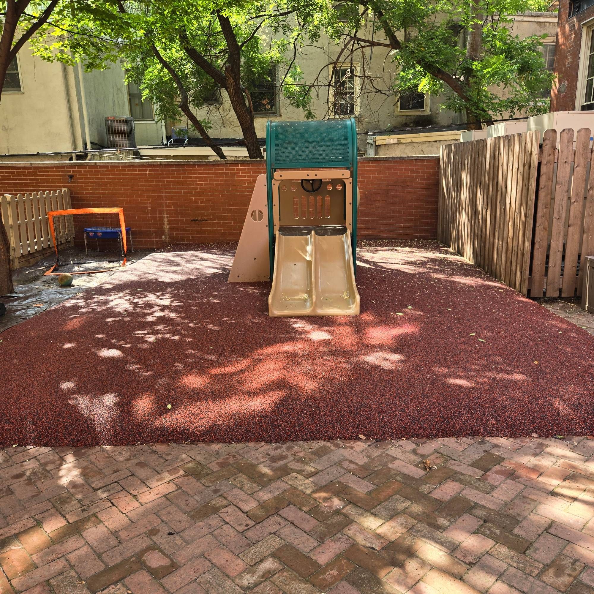 A small double children's slide is in the center of a large red and black rubber mat. It is raised several inches above brick pavers in the yard. On the right is a wood fence. In the rear is a brick wall, behind which are trees and houses.