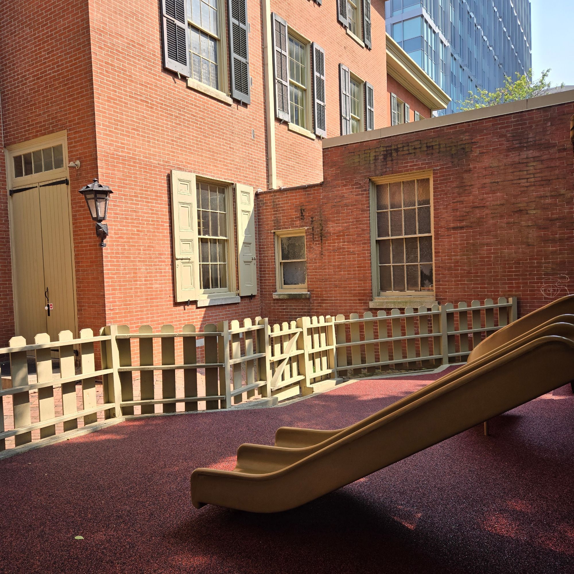 A children's double slide is in the foreground. Around it is a black and red rubber mat, enclosed by a small fence. Behind it is a brick building. It is a Quaker Meetinghouse. The main building has tall doors and windows, with wood shutters next to the windows. In the rear is a modern office building.