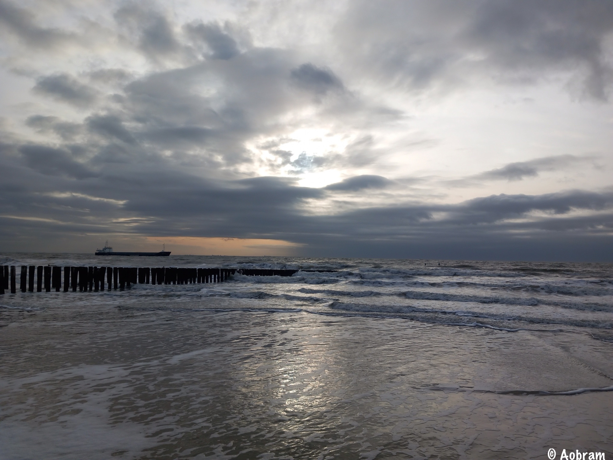 Zonsondergang aan het strand met golfbreker palen en zeeschip aan de horizon.