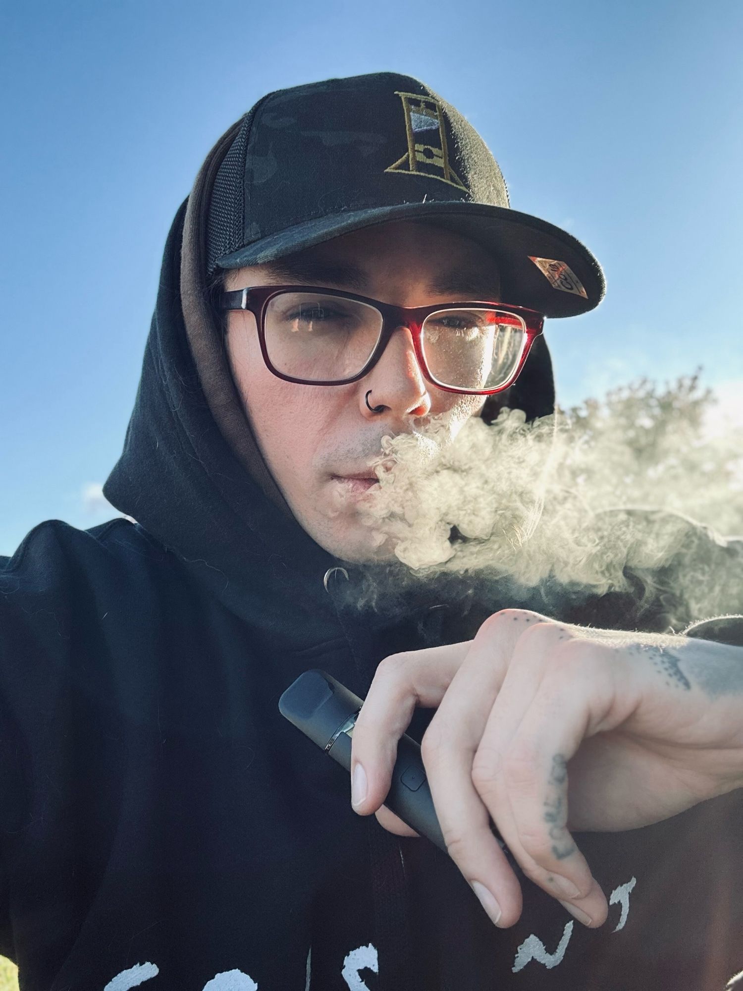 selfie of me in a black hoodie and my cola corporation guillotine hat. you can see my finger tattoo that says forever. I am exhaling vapor from my weed cart