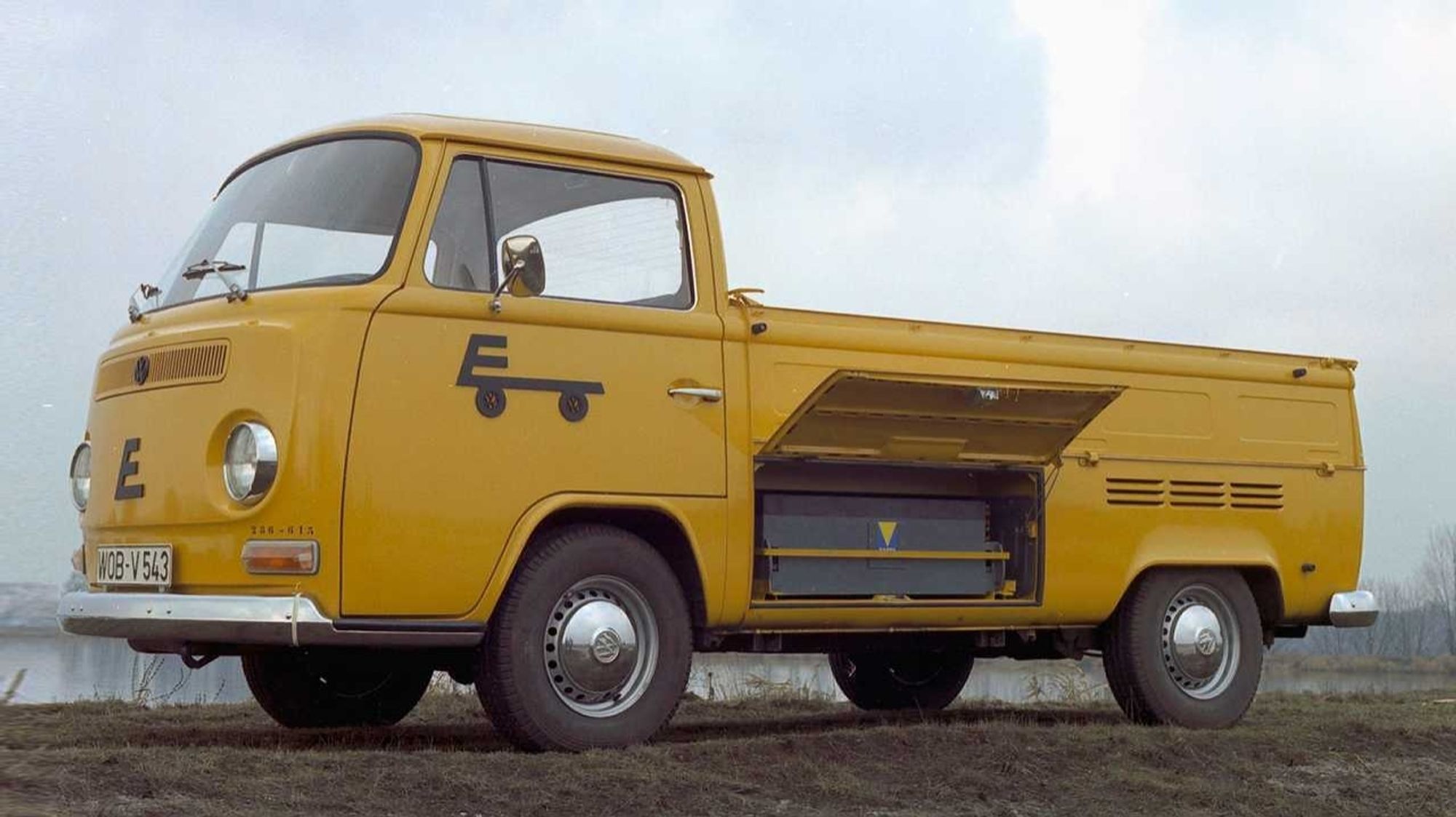 Volkswagen Elektro-Transporter: Optisch serienmäßiger gelborangefarbener VW-Pritschenwagen von 1971, hinter dessen offener Seitenklappe unter der Ladefläche ein riesiger Bleiakku zu erkennen ist.