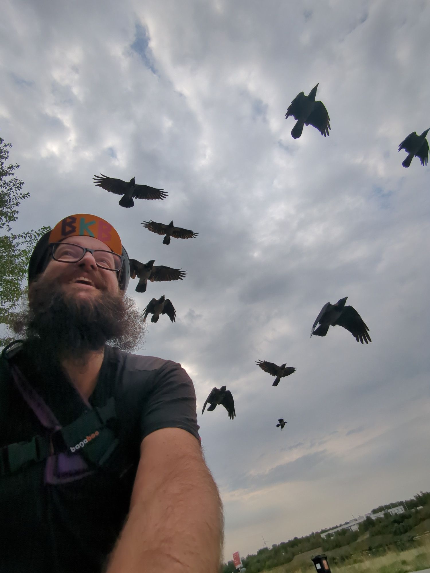 Selfie von mir beim Radeln, ich (Mann mit Vollbart, bunter Kappe, schwarzem Shirt und Brille) bin links lachend im Bild, hinter und über mir fliegen mehrere große dunkle Krähen mit. Der Himmel über uns ist bewölkt und grau.