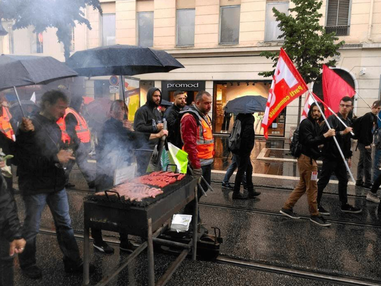 Foto einer Demonstration, hinter Personen mit Flaggen fährt ein Grill voller Würste und Fleisch auf einem speziell angefertigten Gestell auf den Schienen mit.