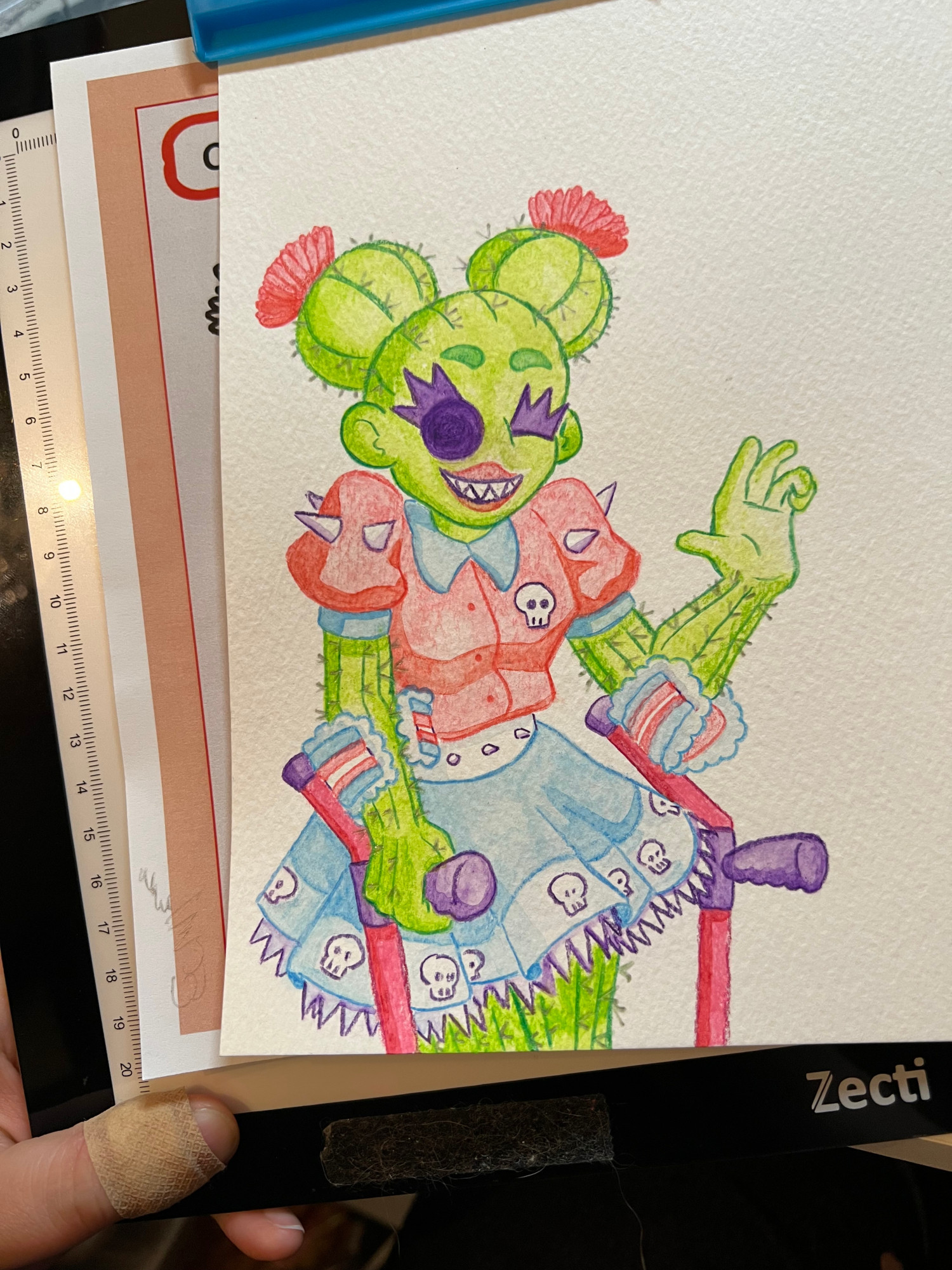 A phone photo of Carmen. She is green with spikes all over her body. Her hair is two ball cactus "buns" with pink flowers growing out of them. She has a pink shirt with a blue collar and puffy short sleeves, with spikes on the shoulder and a skull patch. Her skirt is blue with pointed white frills and a similar skull pattern around the bottom. She uses a pair of forearm crutches with trans flag arm pads.