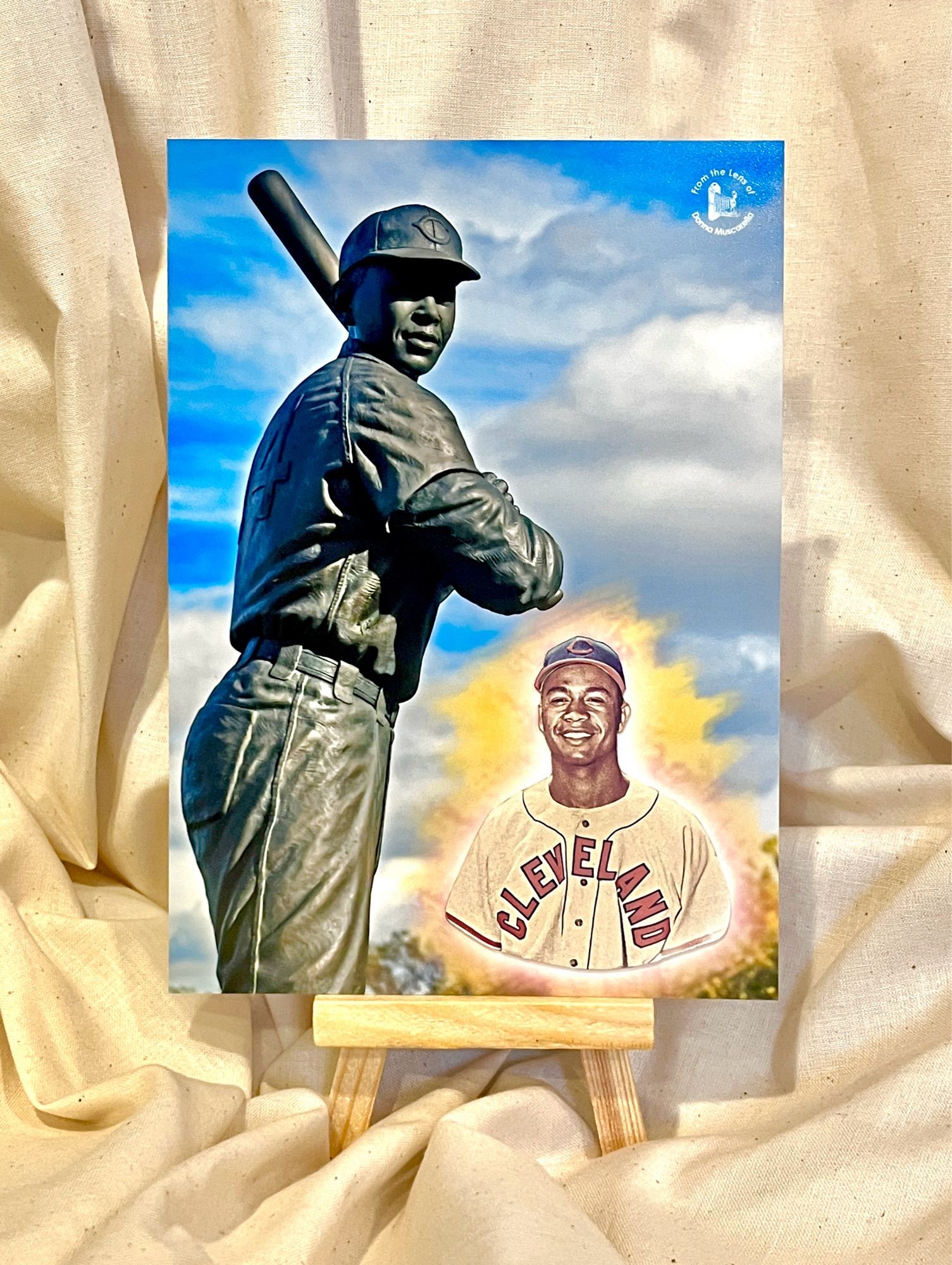 A photograph of a statue of baseball player Larry Doby at bat combined with a portrait of Doby cut from a baseball card. The statue’s backdrop is a bright blue sky with puffy white clouds.